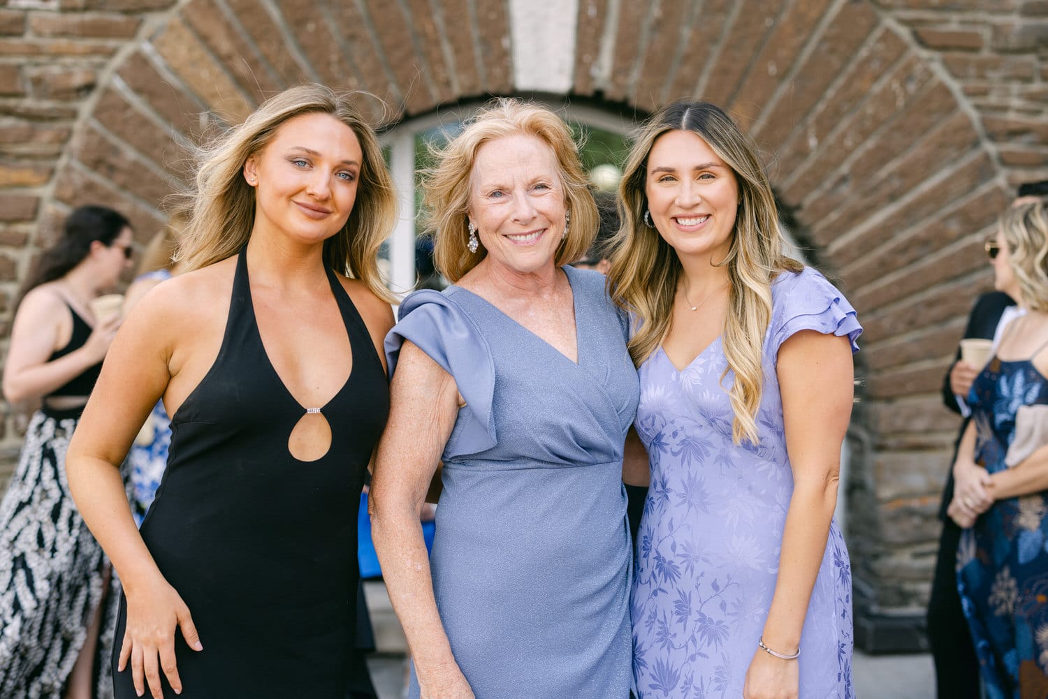 Three women smiling together at a gathering, dressed elegantly against a rustic stone backdrop.