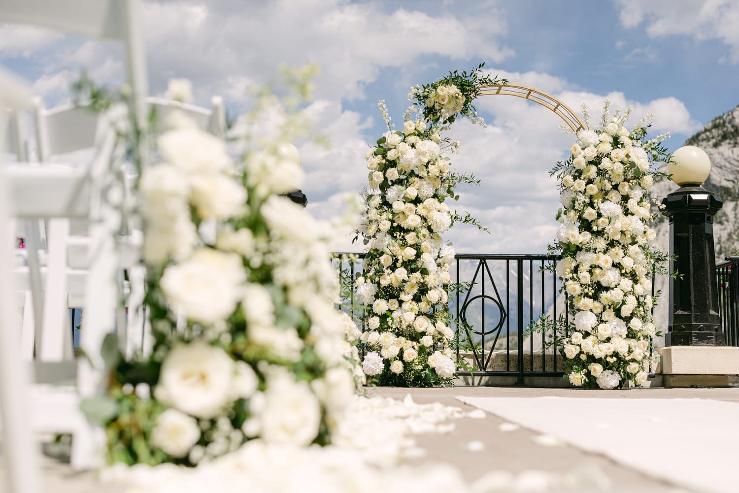 A beautifully arranged floral arch made up of white flowers and greenery, set against a backdrop of clouds and mountains, leading to a stylish outdoor wedding altar.