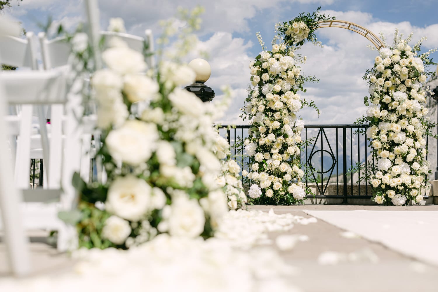 Wedding Floral Archway and Decor::An elegant wedding setup featuring a floral arch adorned with white roses and greenery, surrounded by white chairs and scattered flower petals against a beautiful sky.