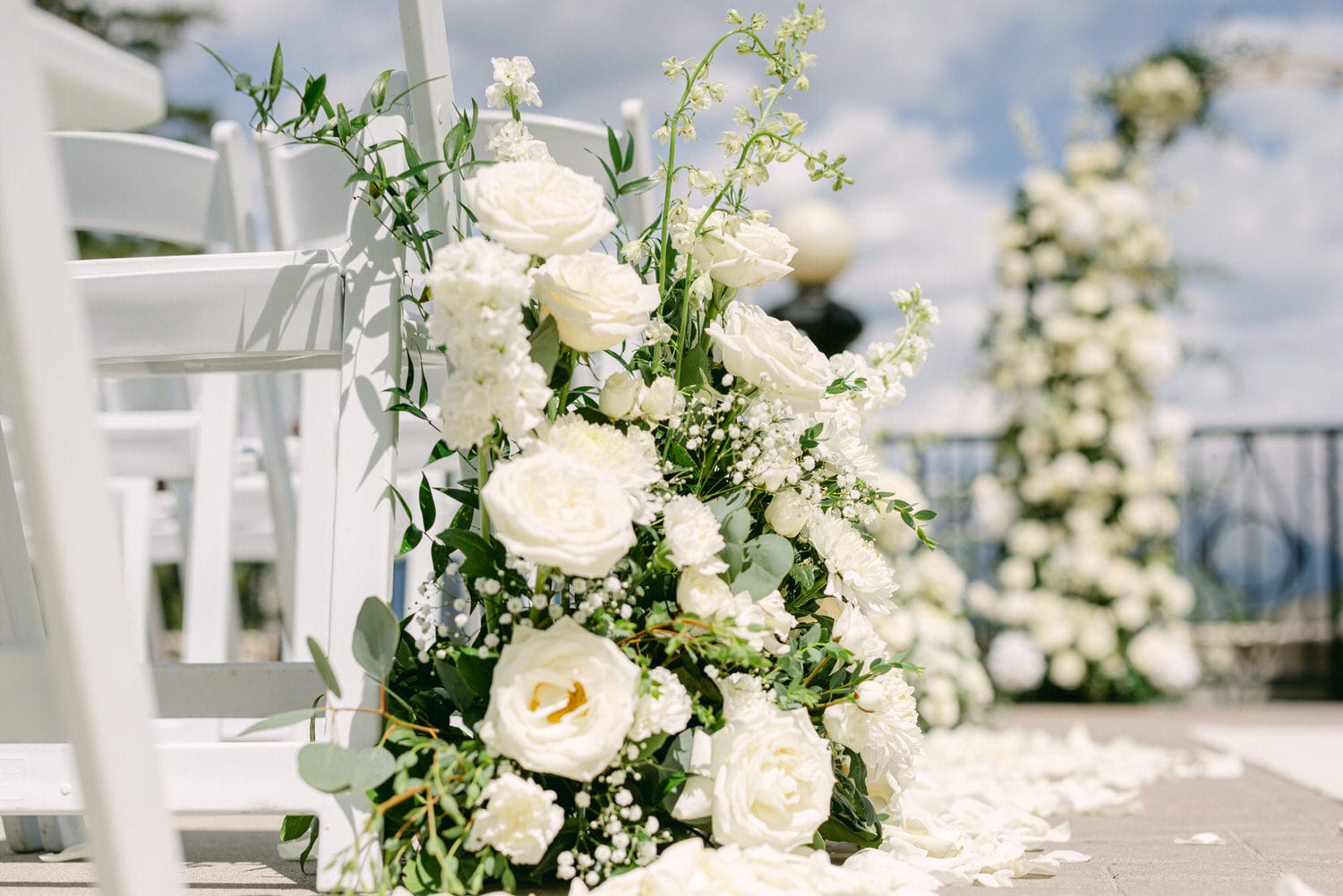 A beautiful arrangement of white roses and greenery adorning a wedding ceremony setup, with white chairs and a floral arch in the background.