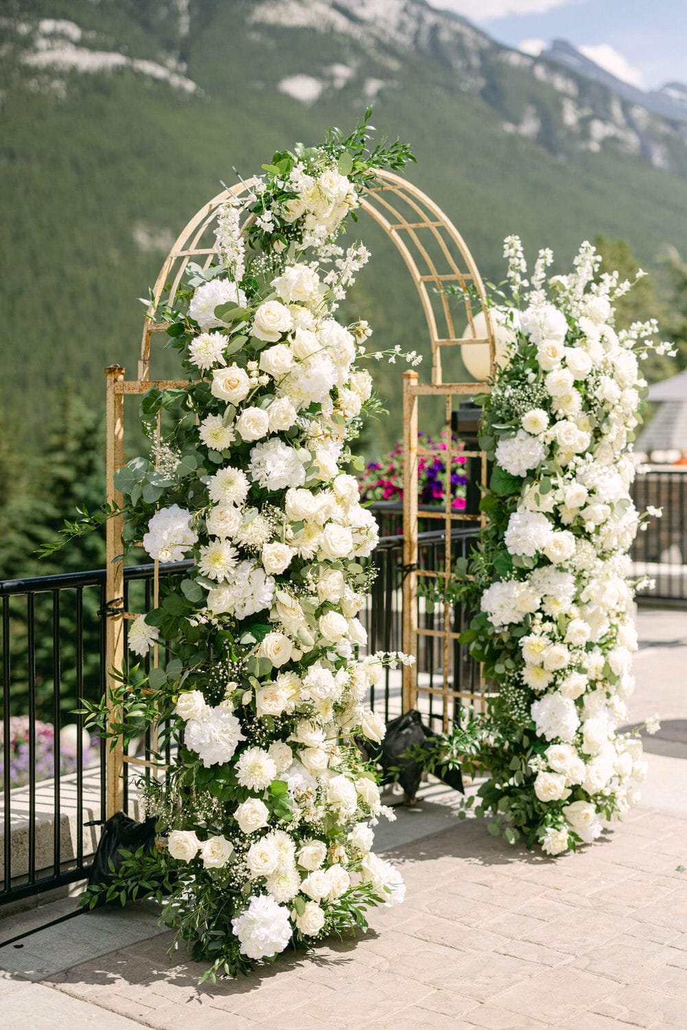 A beautifully decorated wedding arch surrounded by white flowers and greenery, set against a mountainous backdrop.