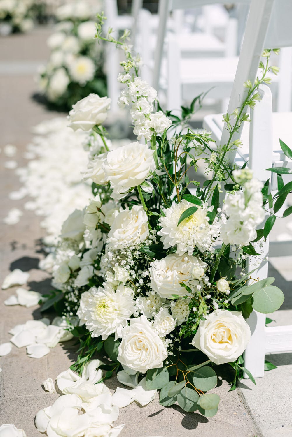 A close-up of a beautiful arrangement of white flowers, including roses and chrysanthemums, accompanied by greenery, set beside white chairs and scattered rose petals on the ground.