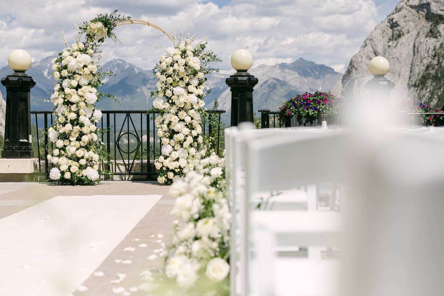 An elegant wedding arch adorned with white flowers set against a stunning mountain backdrop, with rows of white chairs and colorful flower arrangements in the foreground.