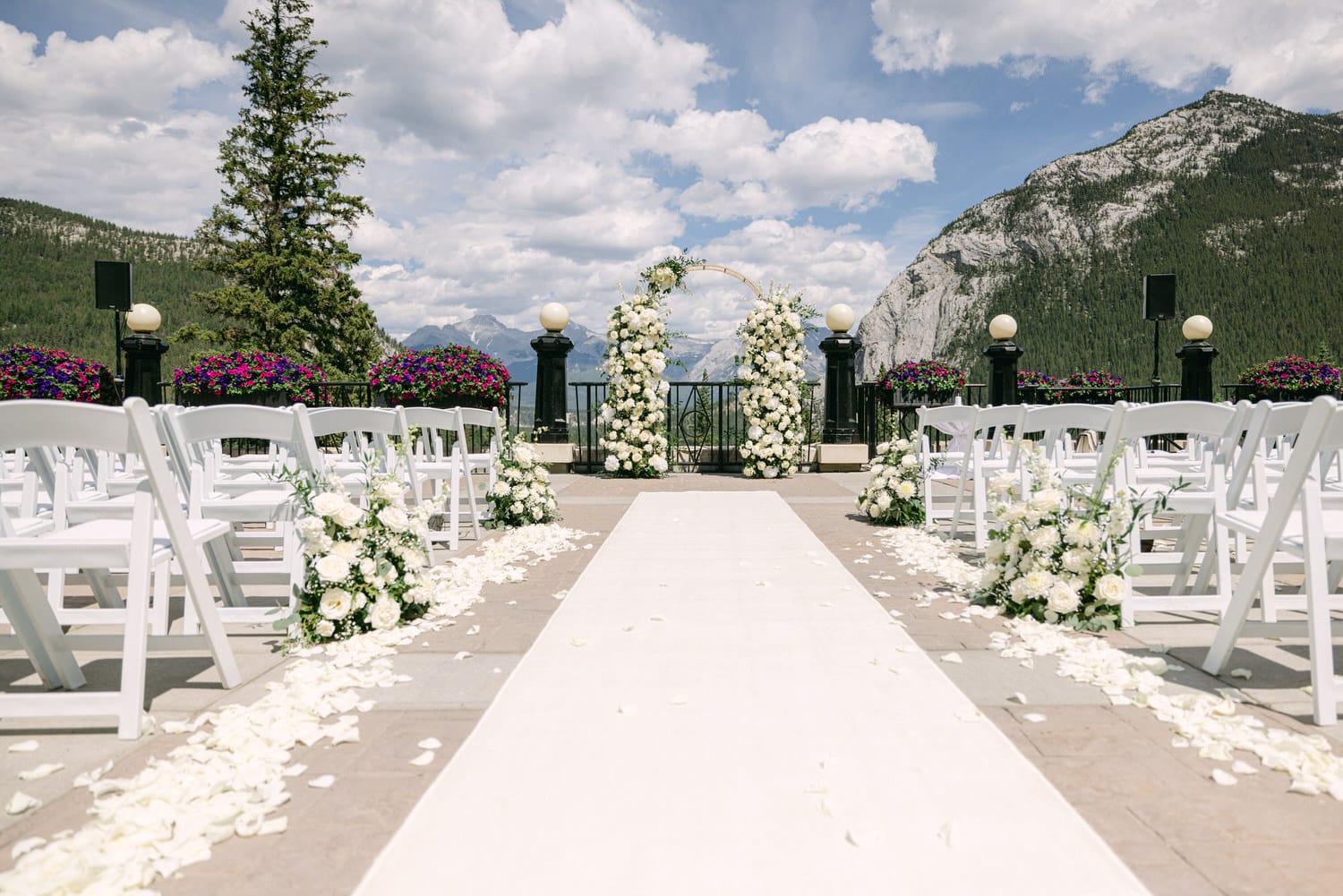 A beautifully arranged outdoor wedding ceremony with white chairs, floral decorations, and a stunning mountain backdrop under a partly cloudy sky.