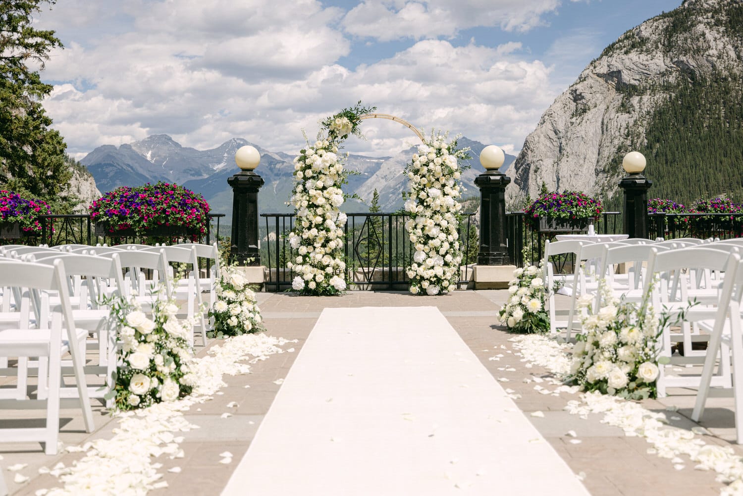 An elegant outdoor wedding setup featuring white floral arrangements, a flower-adorned arch, and a scenic mountain backdrop with chairs arranged along a petal-strewn aisle.