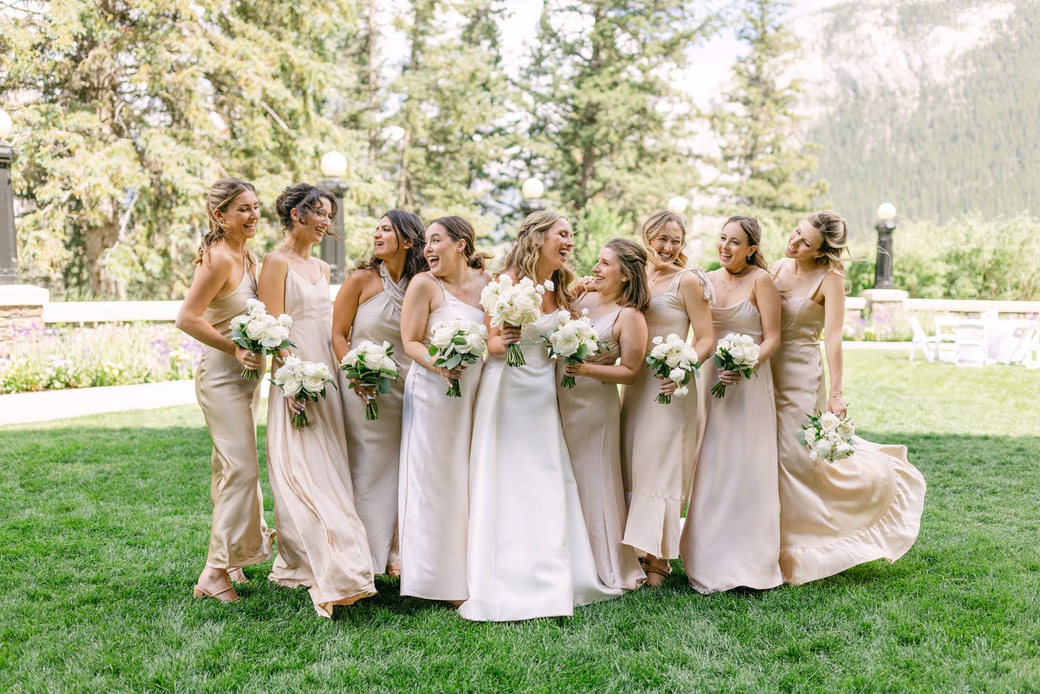 A joyful group of bridesmaids dressed in elegant beige gowns holding white bouquets, posing together in a lush green outdoor setting.