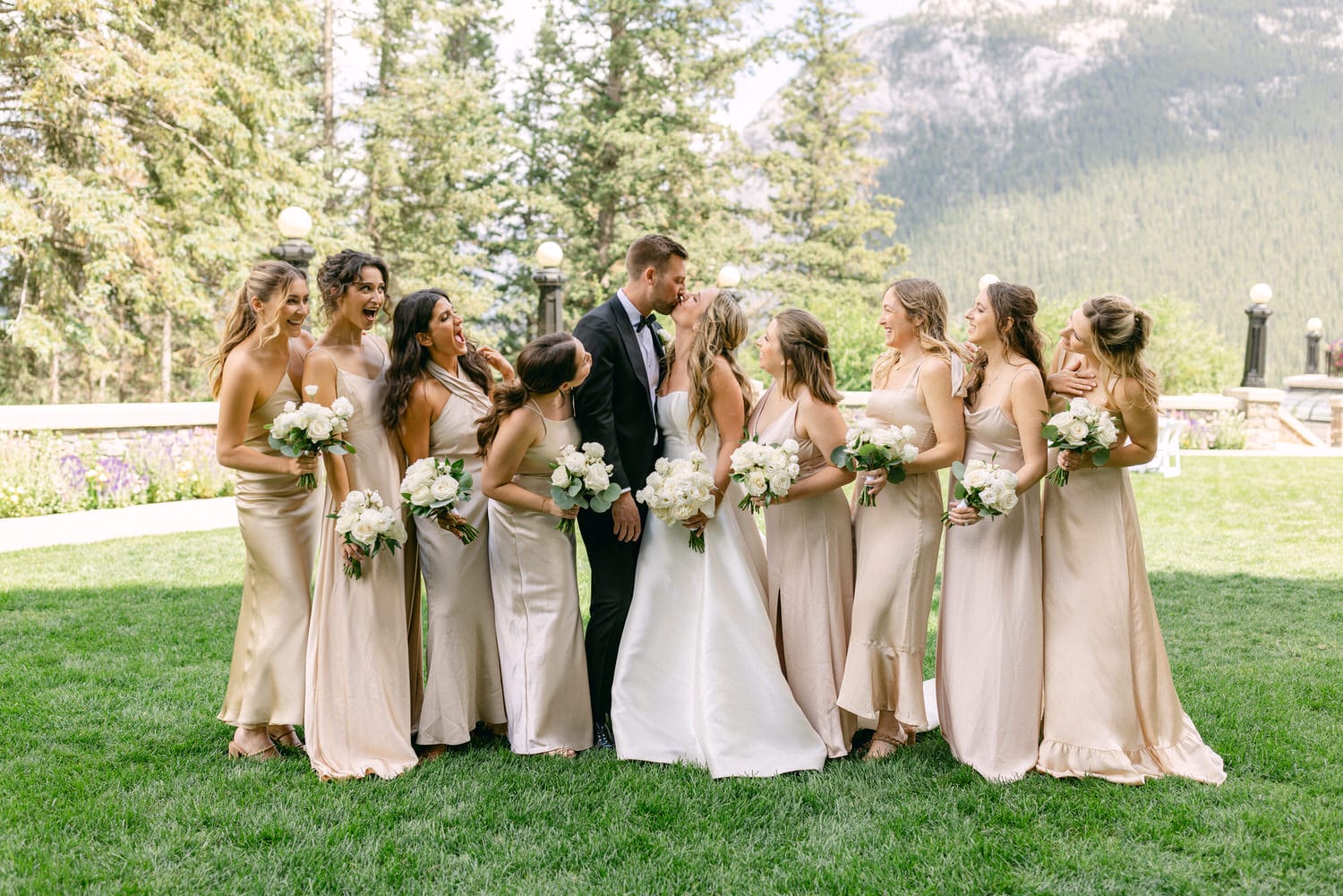 A joyful couple sharing a kiss surrounded by their bridesmaids, all dressed in elegant attire, set against a stunning natural backdrop.