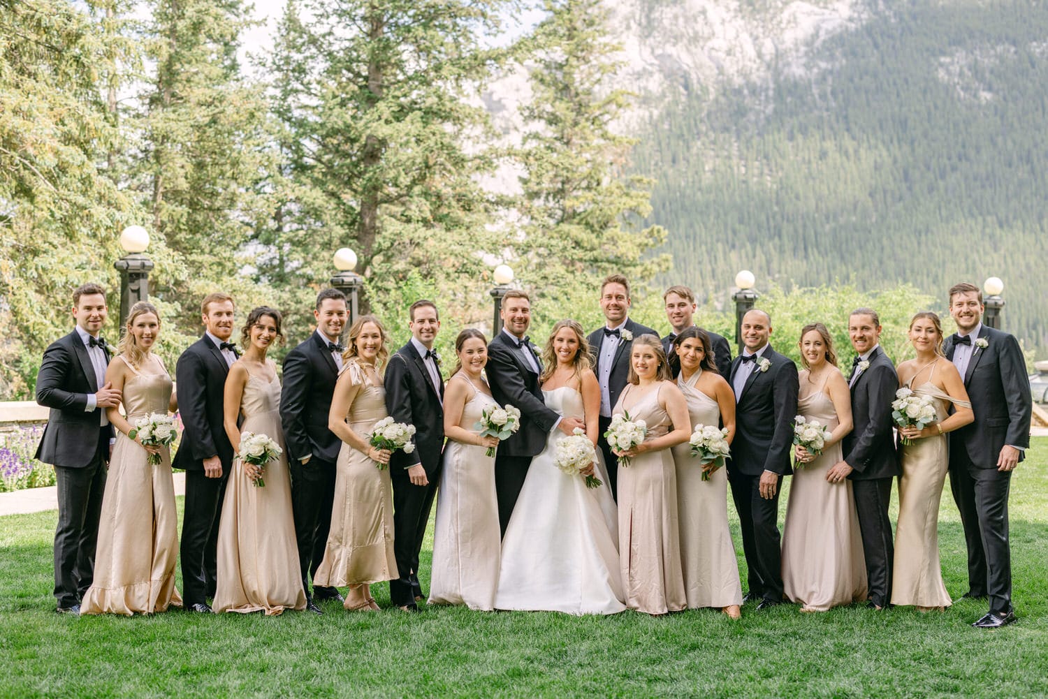 A joyful wedding party poses elegantly outdoors, dressed in formal attire with a mountainous backdrop and lush greenery, capturing a moment of celebration.