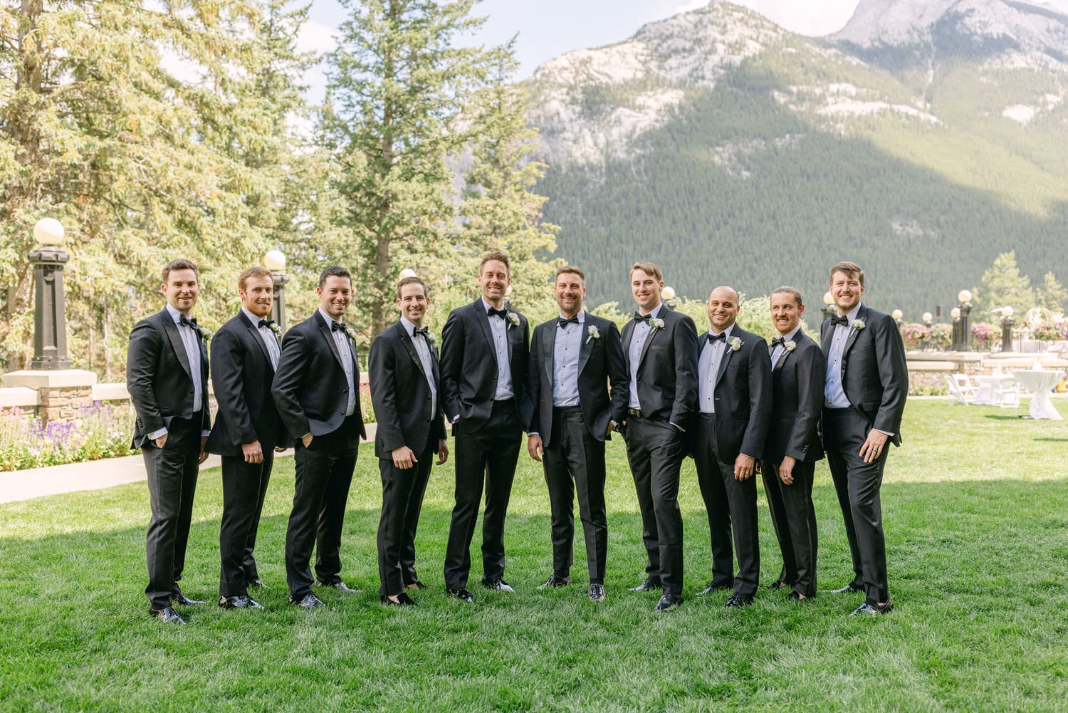 A group of ten men in tuxedos and bow ties, posing outdoors against a scenic backdrop of trees and mountains.