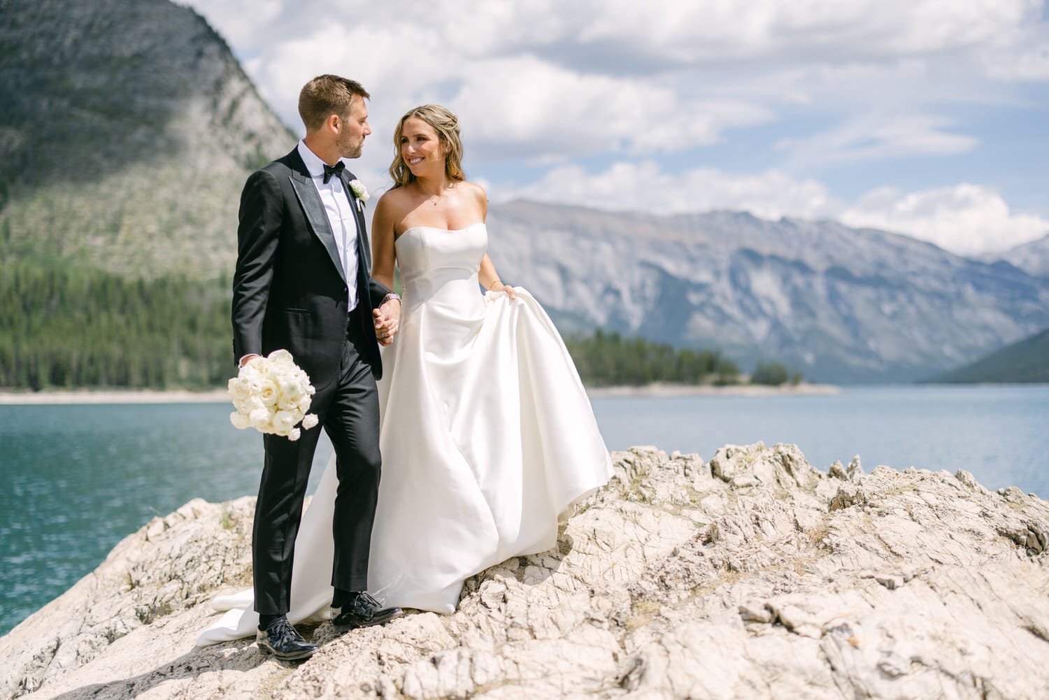 A happy couple in formal attire stands hand in hand on a rocky shore, with a serene lake and mountains in the background.