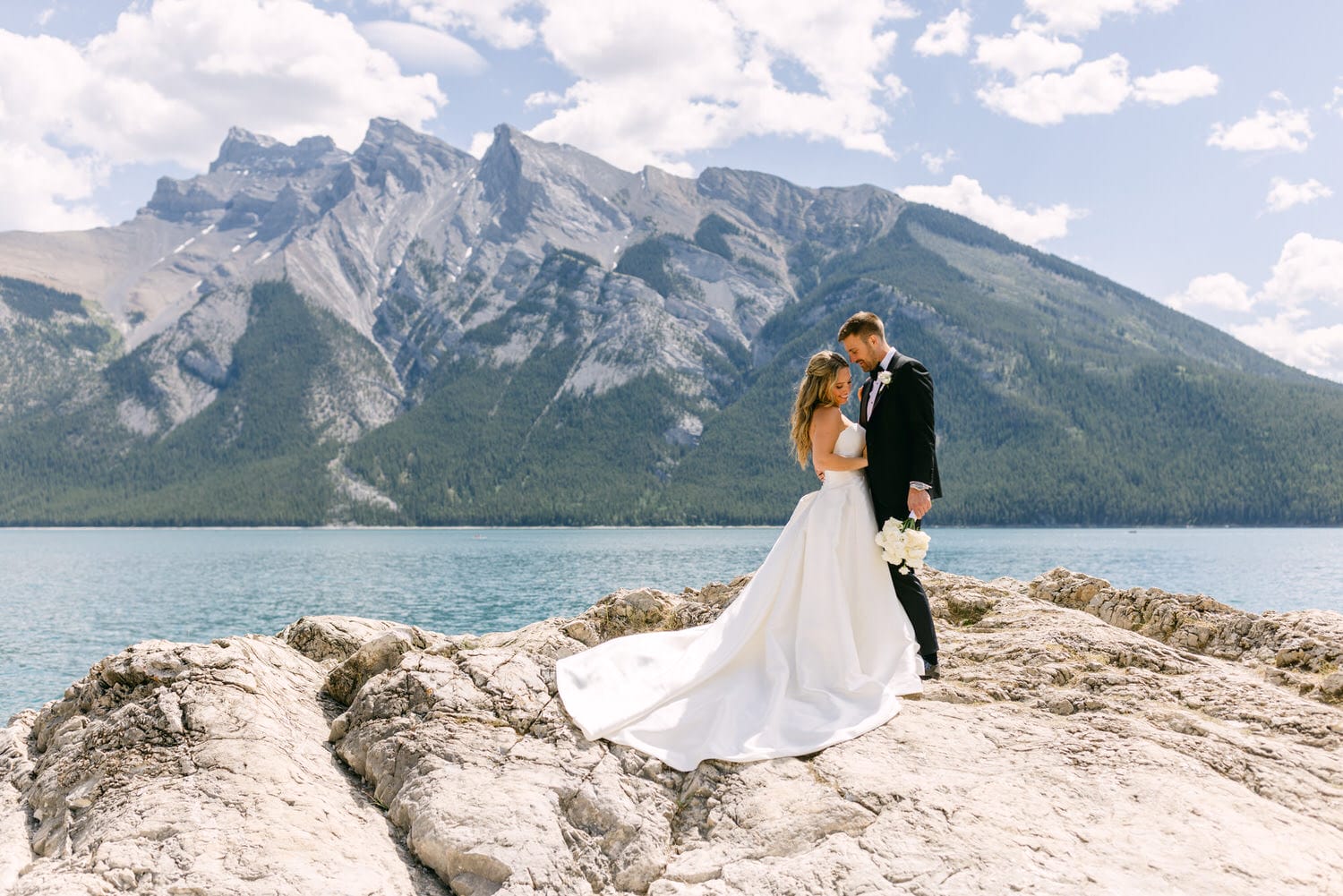 A couple shares a romantic moment on a rocky outcrop beside a serene lake, with majestic mountains in the background.