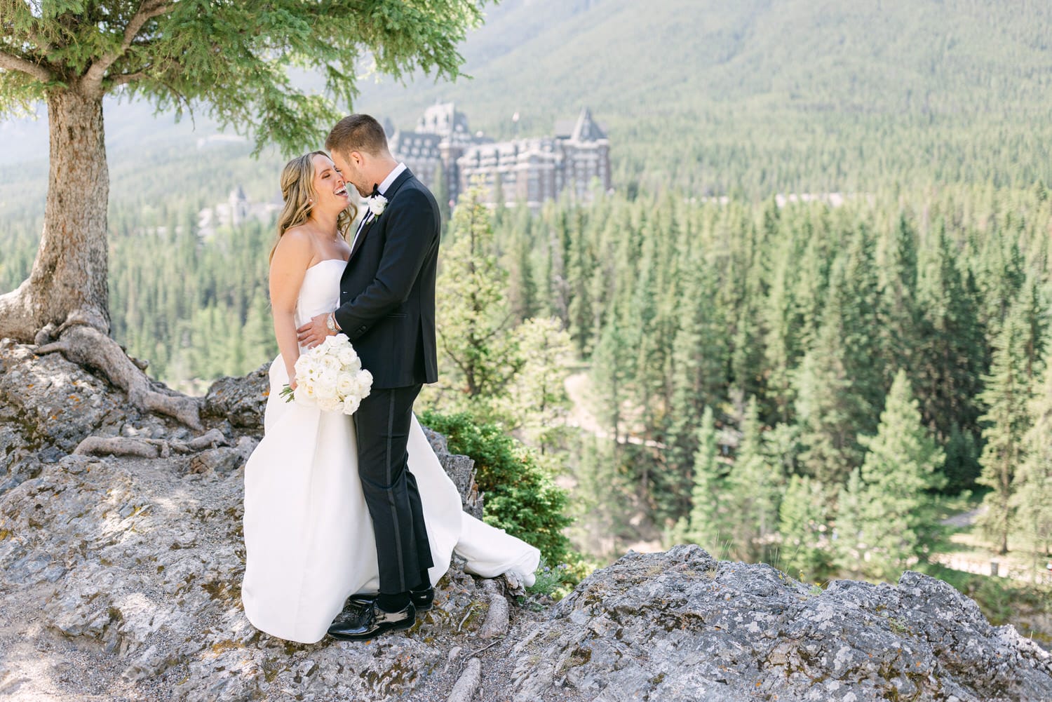 A couple shares a tender moment on a rocky outcrop with a lush green forest and a grand hotel in the background.