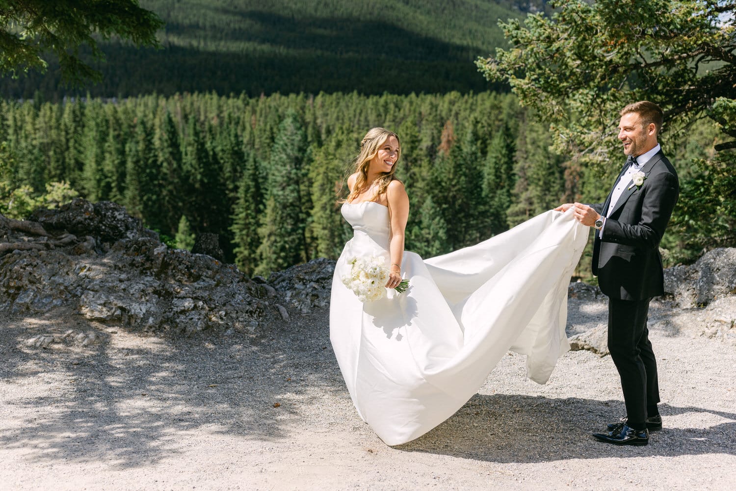 A joyful bride in a flowing white gown and a groom in formal attire pose playfully against a backdrop of lush green trees.