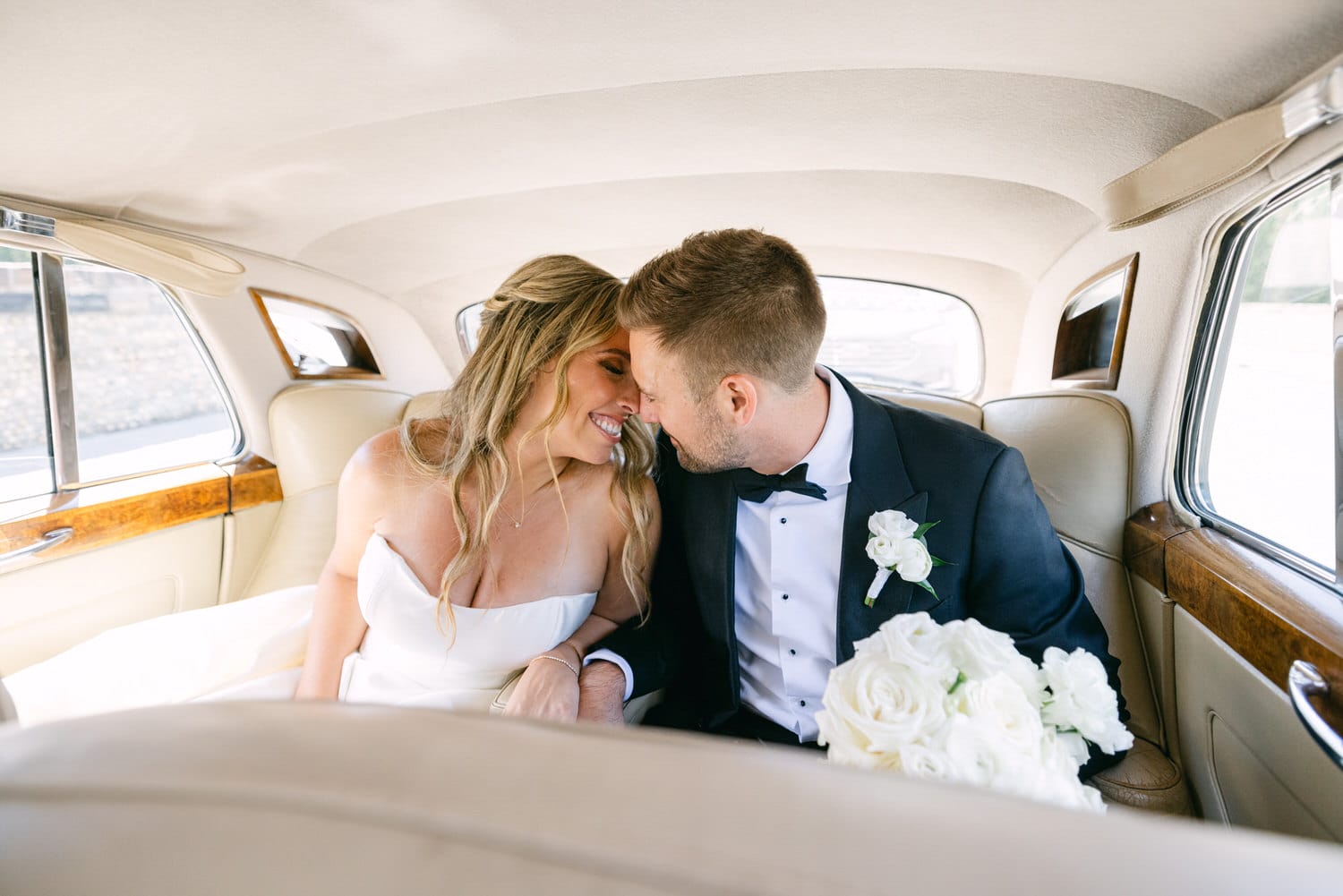 A newlywed couple sharing a joyful smile while nestled together in the back seat of a classic car, surrounded by elegant wedding attire and flowers.