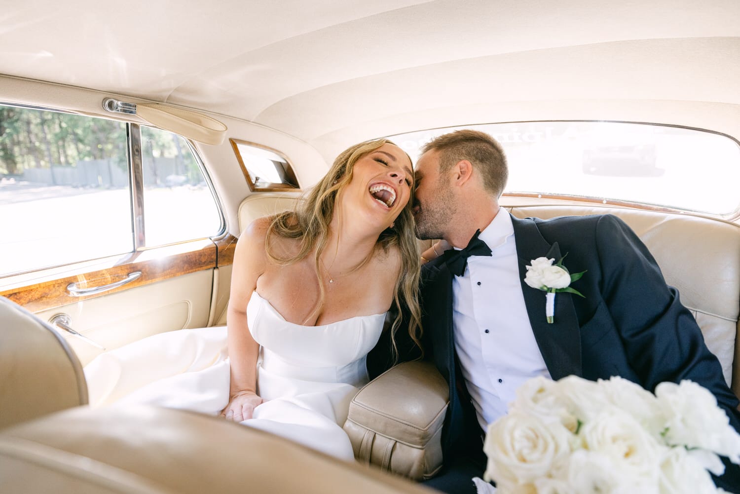 A happy bride and groom share a tender moment in a vintage car, with the bride laughing and the groom whispering affectionately.