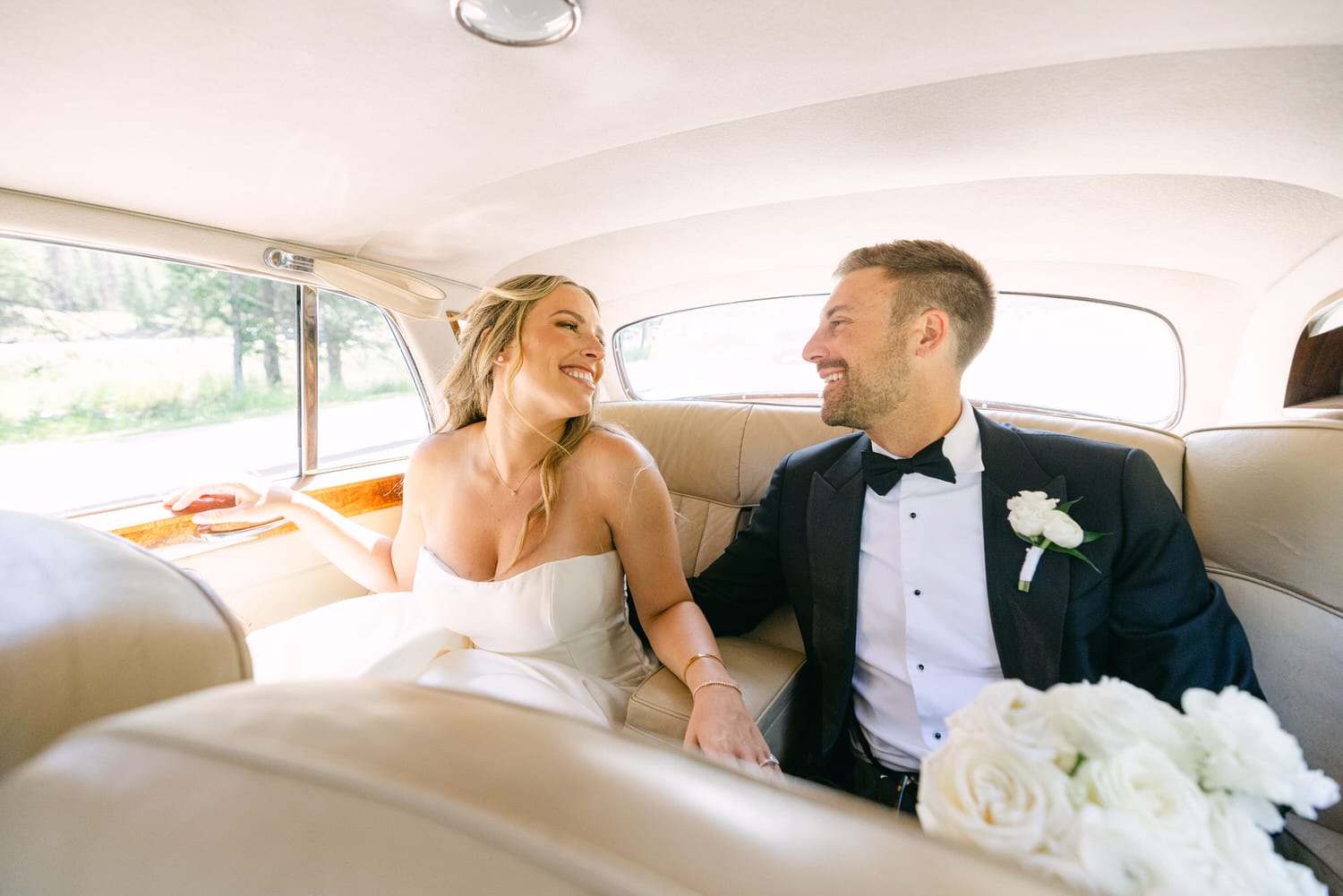 A bride and groom sharing a joyful smile while seated in the back of a vintage car, surrounded by elegant upholstery and natural light.