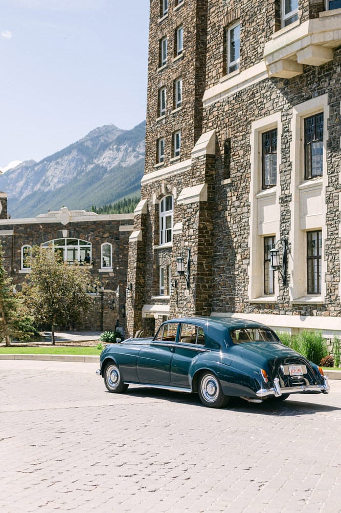 A vintage car parked in front of a stone-clad hotel, surrounded by mountains and greenery.