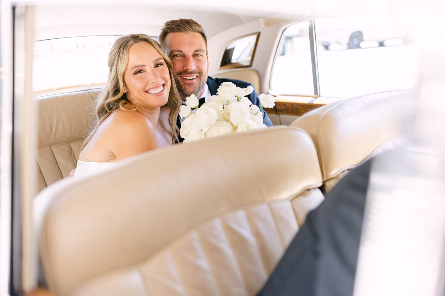 A happy bride and groom embrace in the back seat of a luxurious car, smiling and holding a bouquet of white roses.