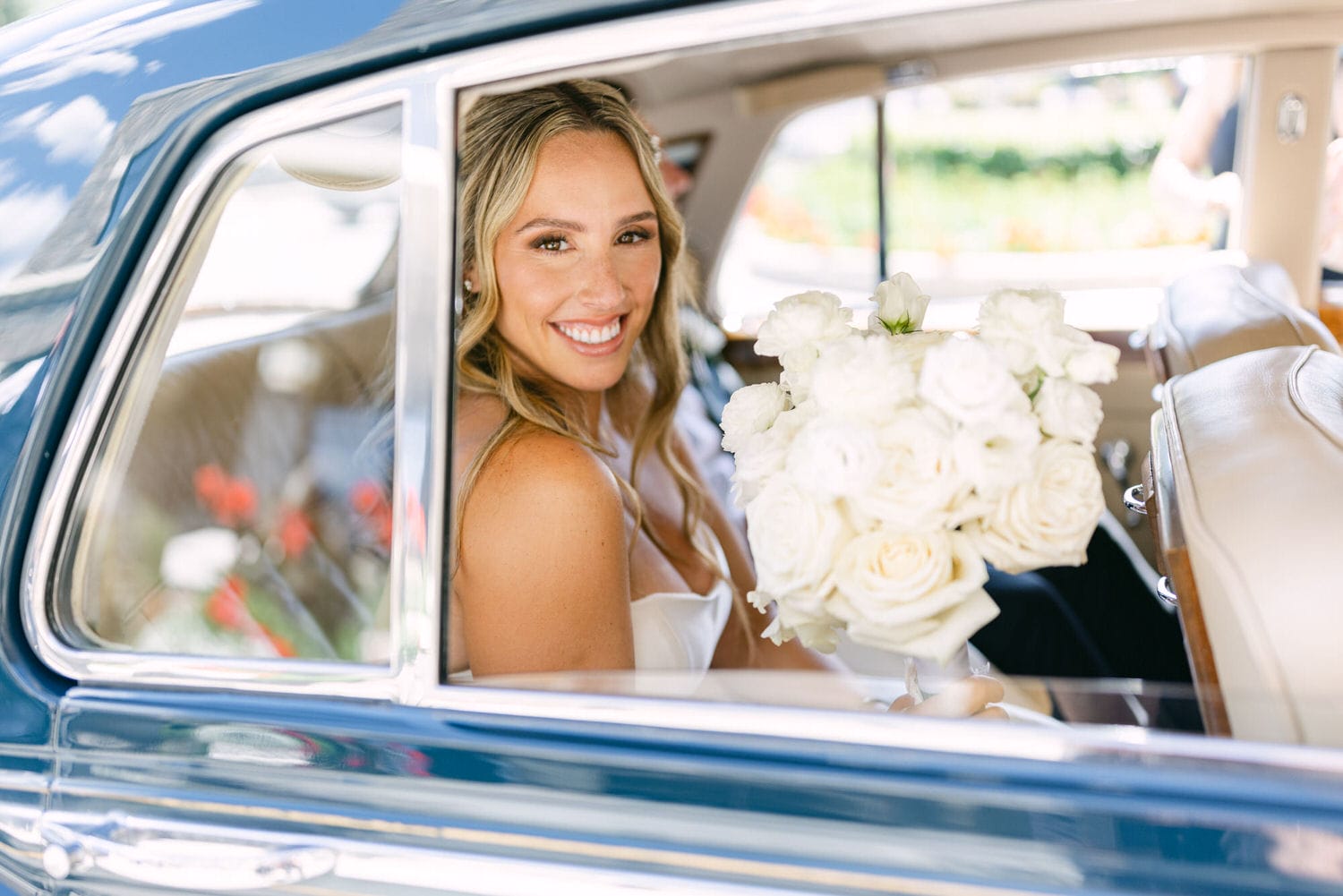 A smiling bride holds a bouquet of white roses while seated in a vintage car, capturing a moment of happiness before her wedding.