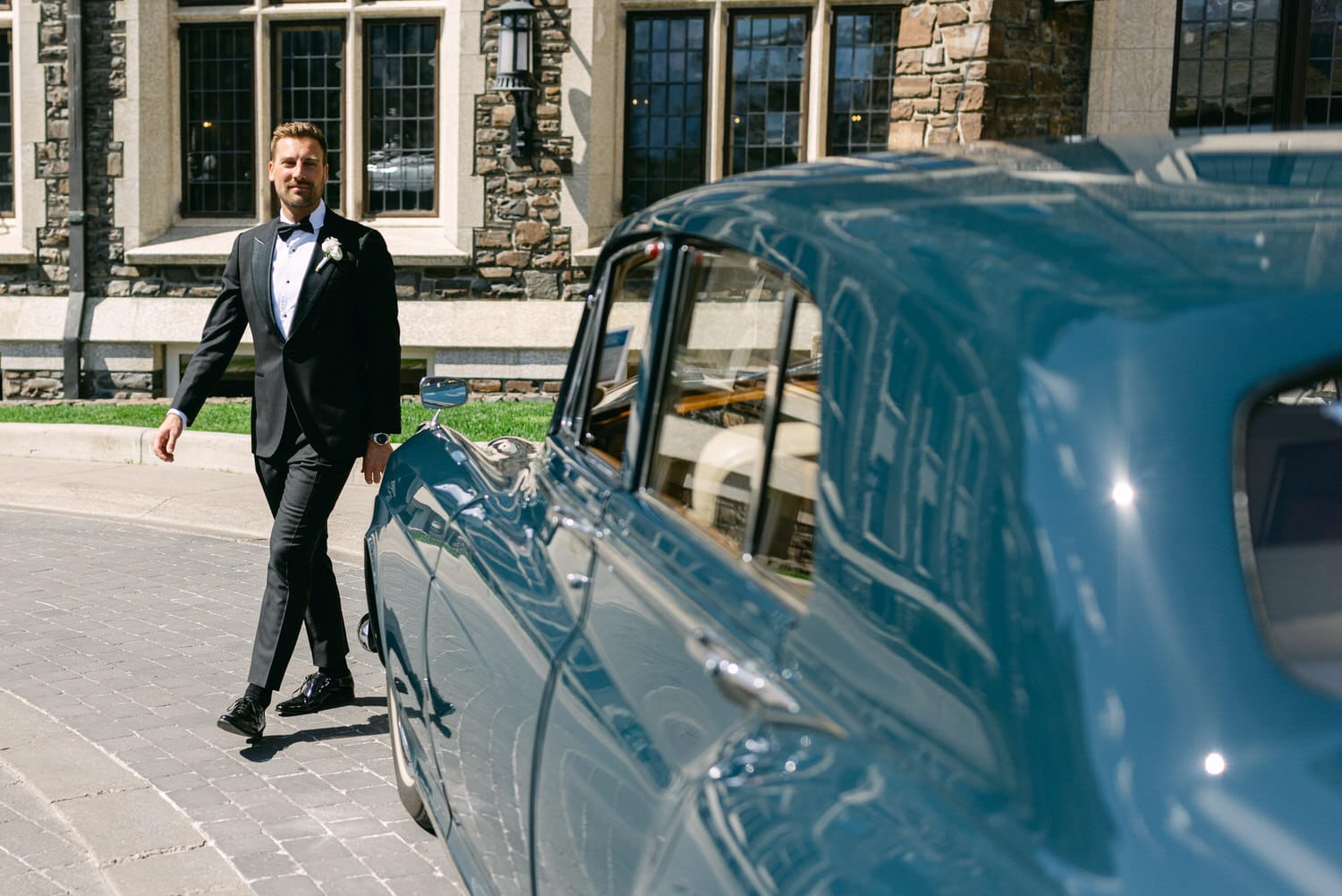 A well-dressed man in a tuxedo walking beside a vintage car in front of a beautiful stone building.