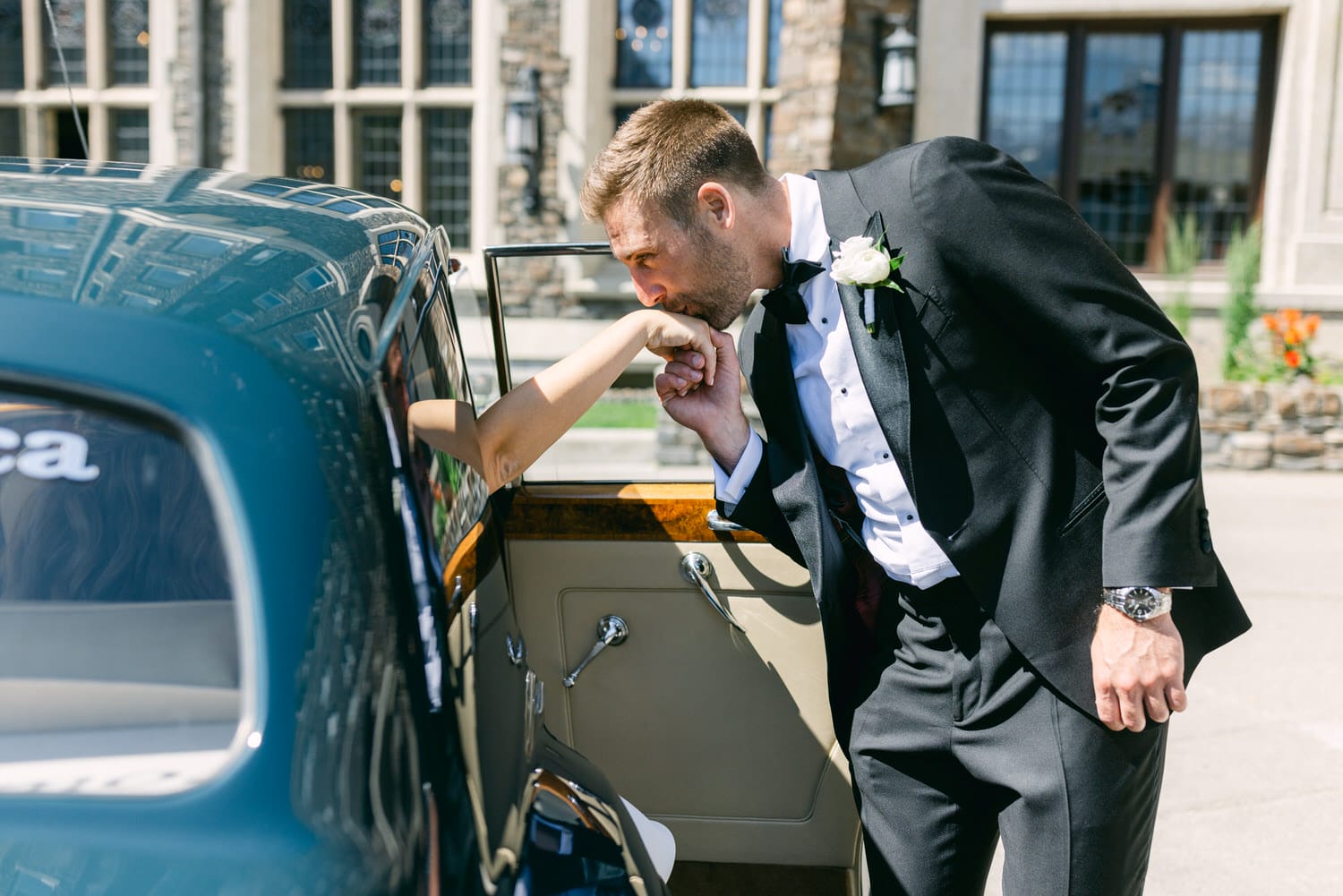 A man in formal attire kissing the hand of a woman reaching out from a classic car, set against an upscale venue backdrop.