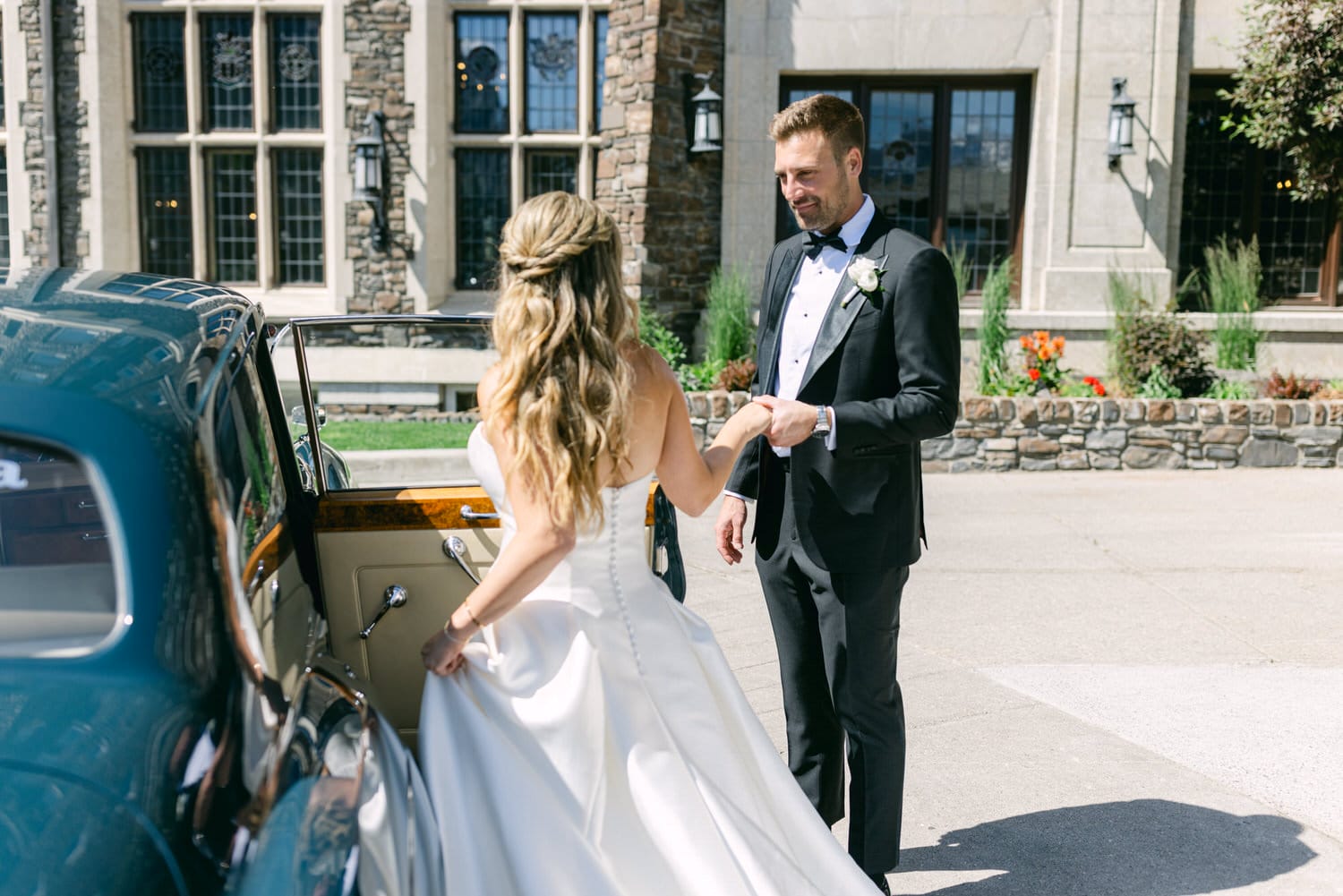 A bride in a flowing white gown is assisted by a groom in a tuxedo as she prepares to enter a classic car outside a beautiful stone building.