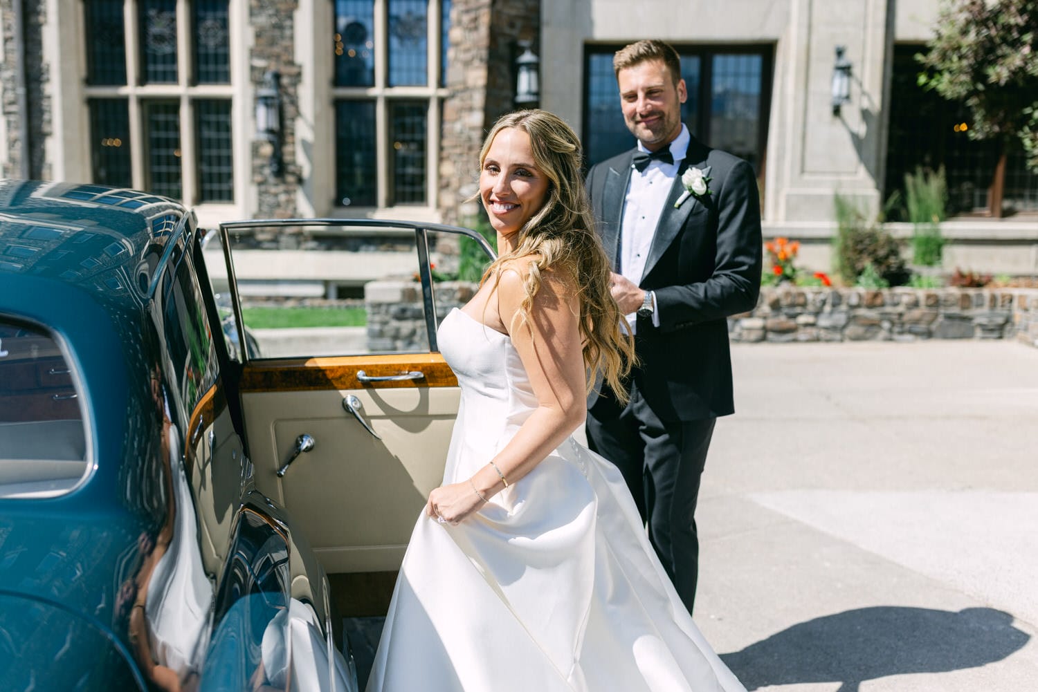 A bride in a beautiful white dress smiles as she prepares to enter a vintage car, while a man in a tuxedo assists her.