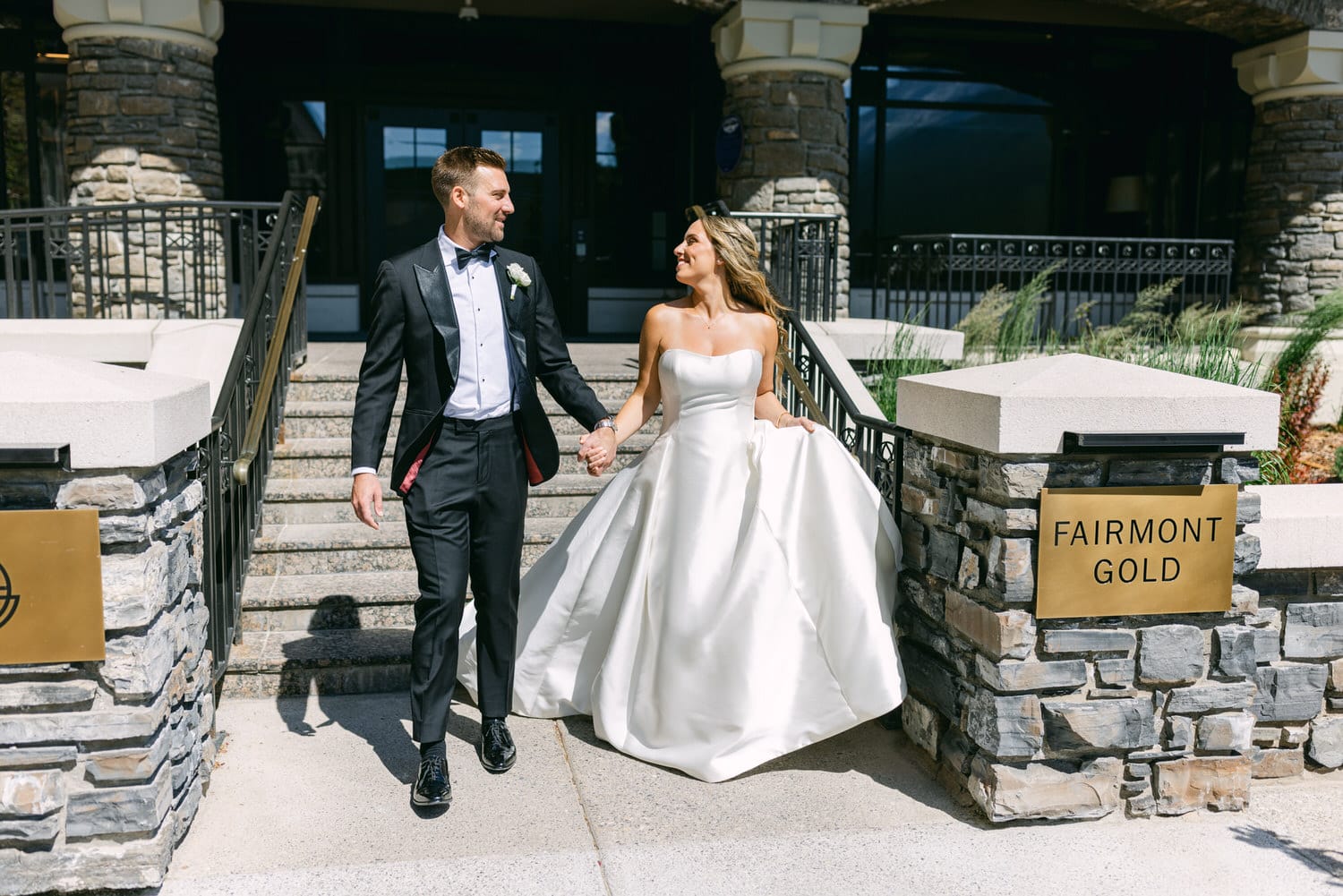 A bride in a white gown and a groom in a tuxedo hold hands while descending steps outside the Fairmont Gold venue, radiating joy and love.