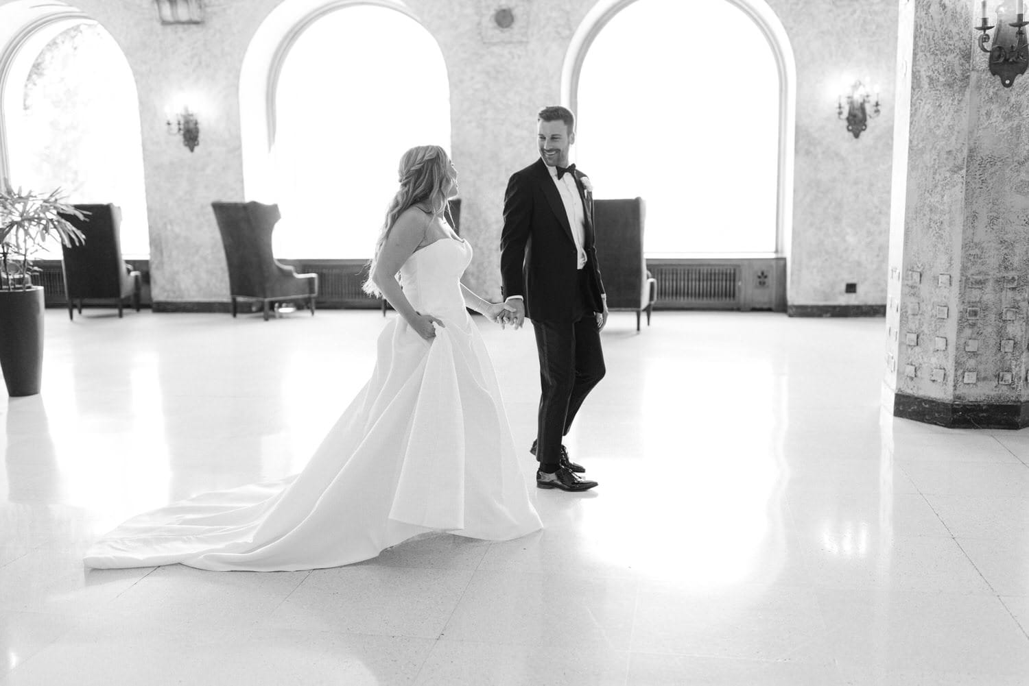 A bride and groom holding hands while walking in an elegant, spacious venue with large windows and stylish furniture.