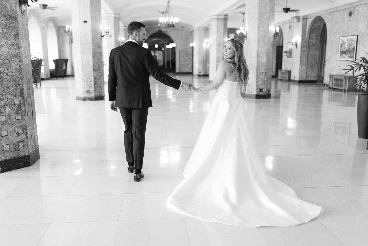 A couple walks hand in hand down a stylish corridor, the bride in a flowing white gown and the groom in a classic black suit, conveying love and joy.