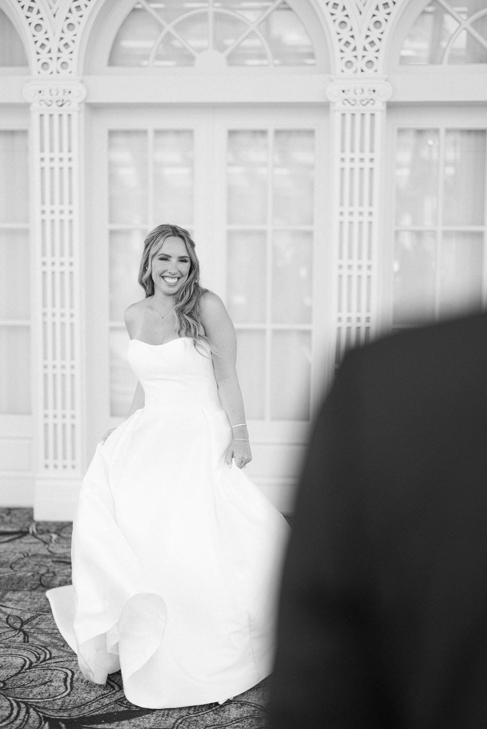 A smiling bride in a strapless white wedding dress enjoys a moment before her partner, captured in a stylish black and white photograph.