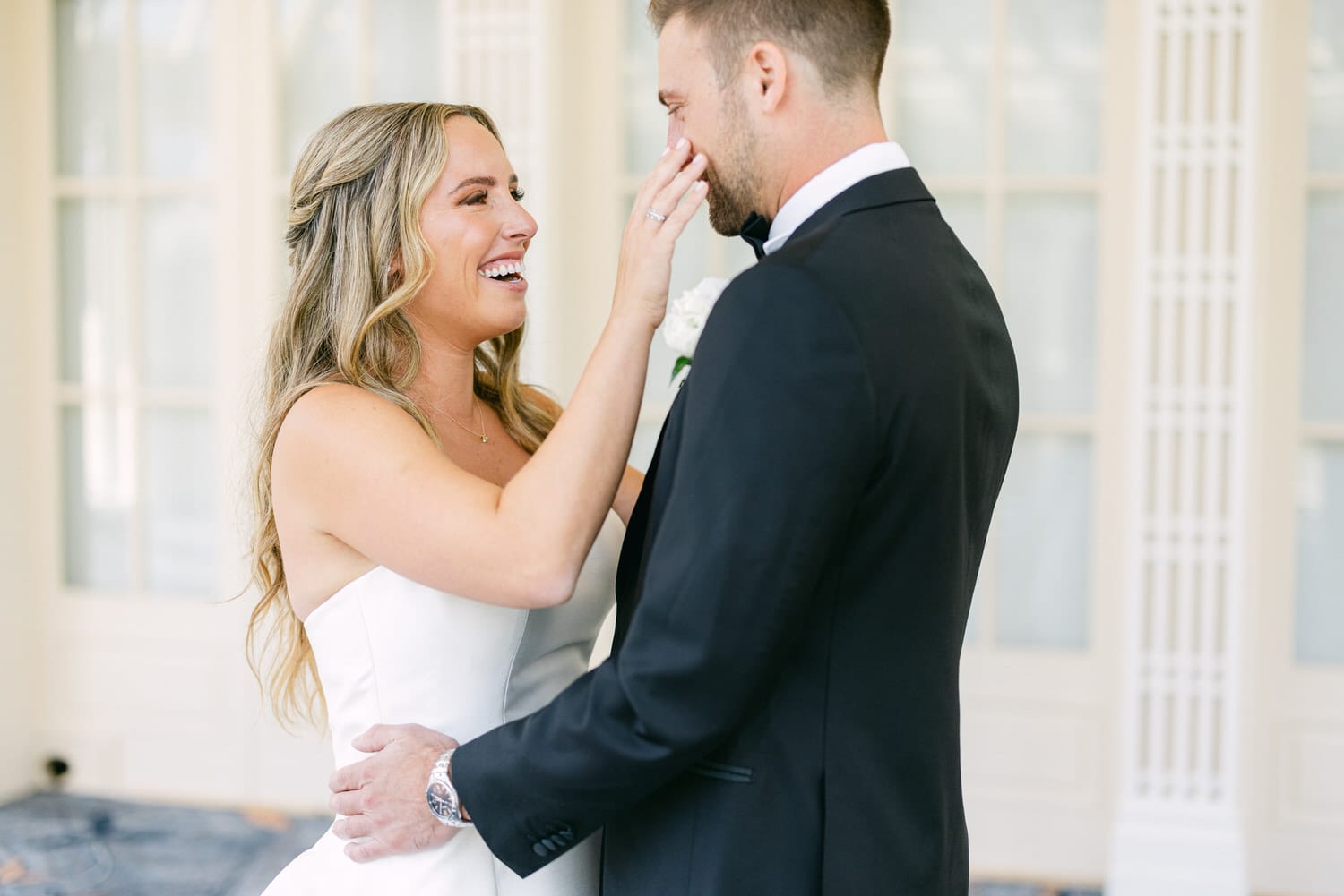 A bride lovingly touches her groom's face, both smiling happily, in a beautifully lit venue.