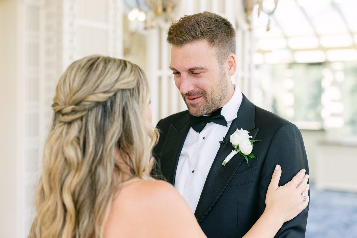 A groom in a tuxedo shares a tender gaze with his bride, who has long, wavy hair, in a beautifully lit setting adorned with elegant decor.