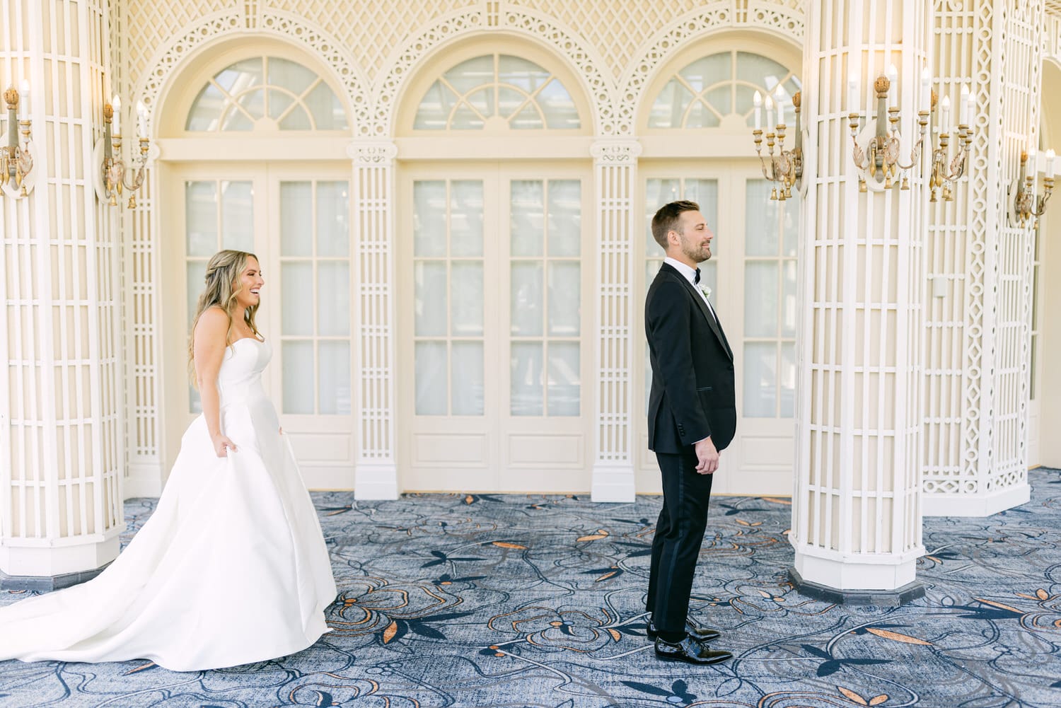 A bride in a stunning white gown smiles at a groom in a tuxedo, both standing in a beautifully designed venue with arched windows and intricate decor.