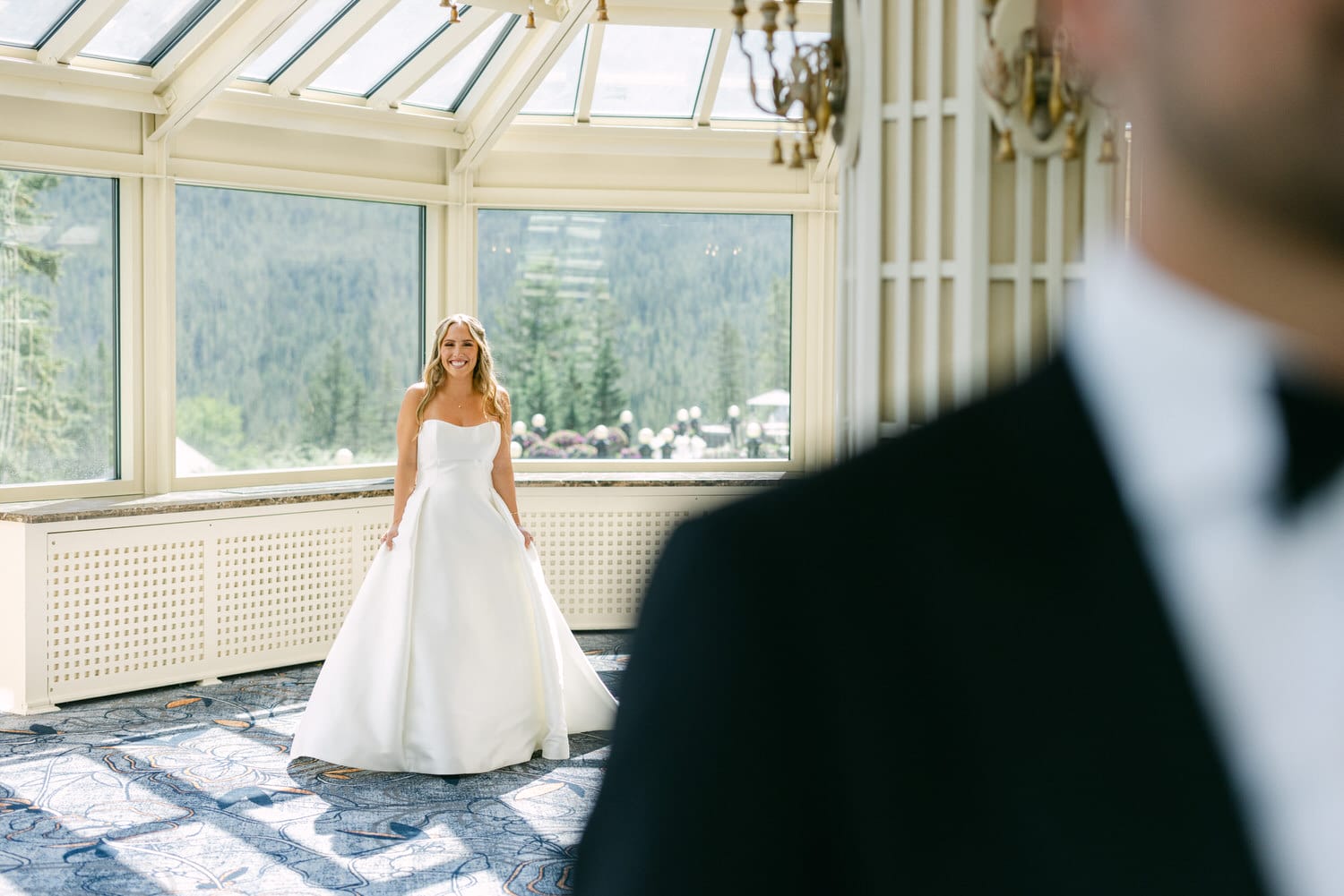 A bride smiles in a beautiful wedding gown, standing elegantly in a sunlit room with panoramic views of nature, as a groom approaches from the side.