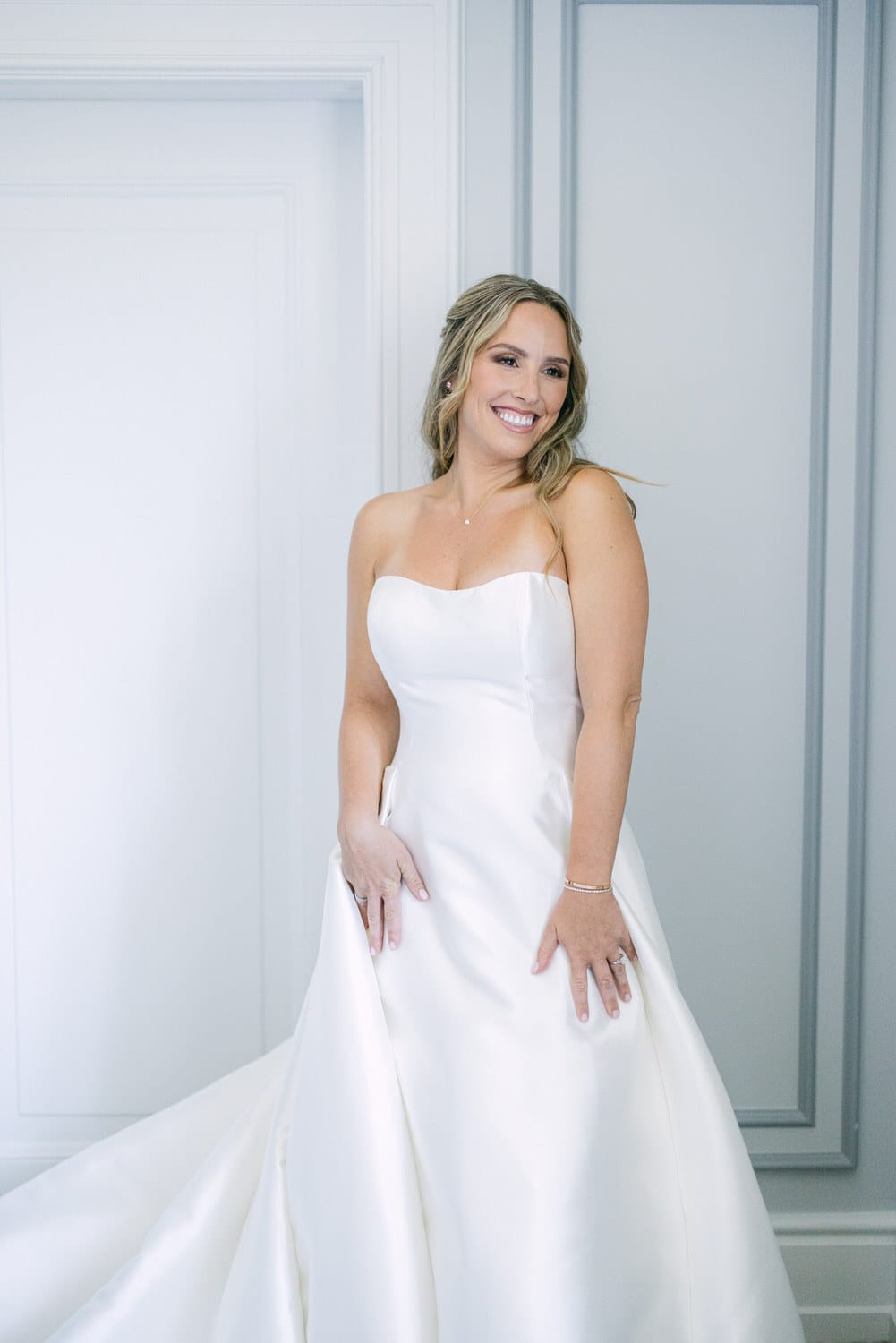 A smiling bride in a strapless white gown stands elegantly against a light backdrop, showcasing her joyful demeanor.