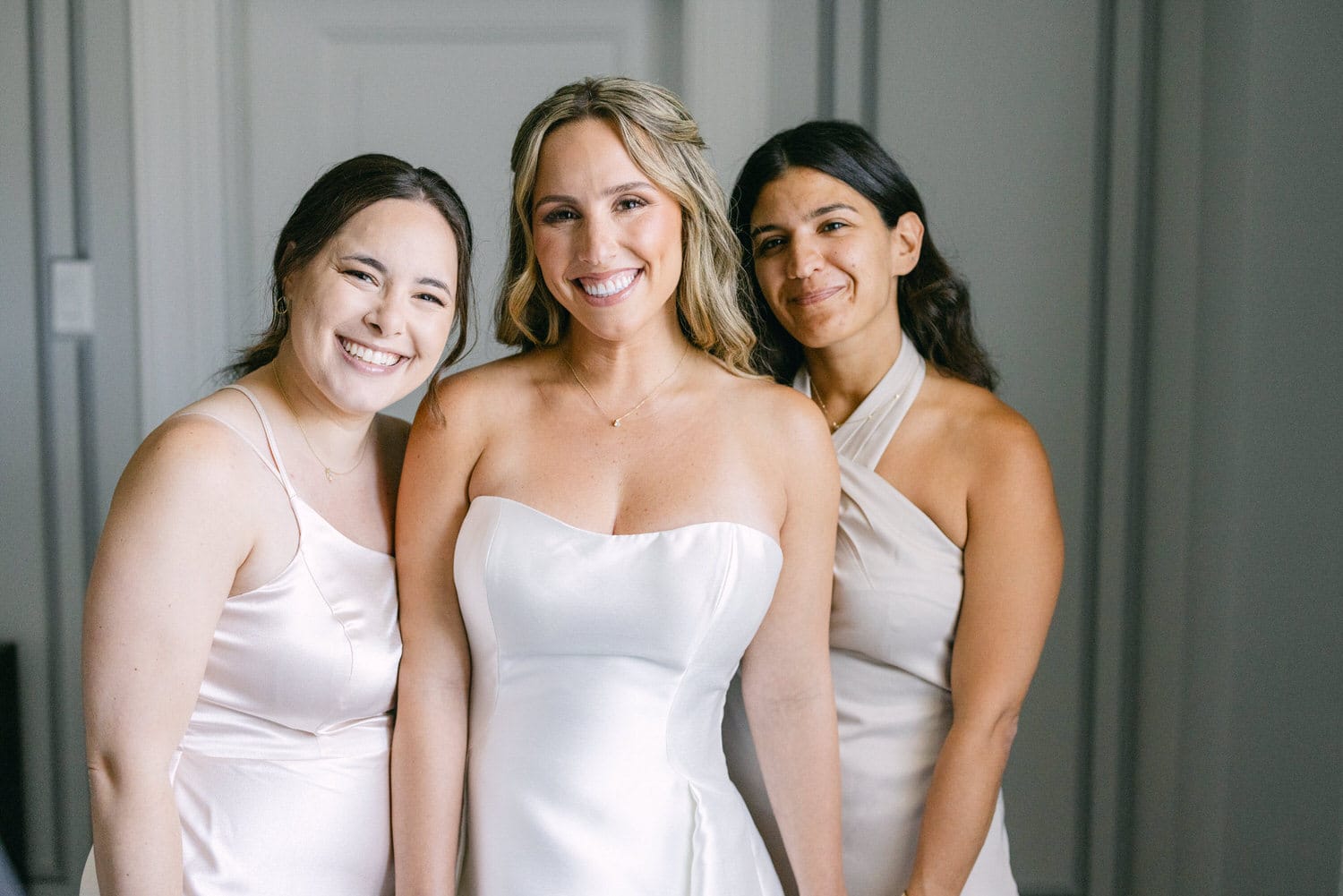 Three women in elegant dresses pose happily together, capturing a joyful moment before a wedding.