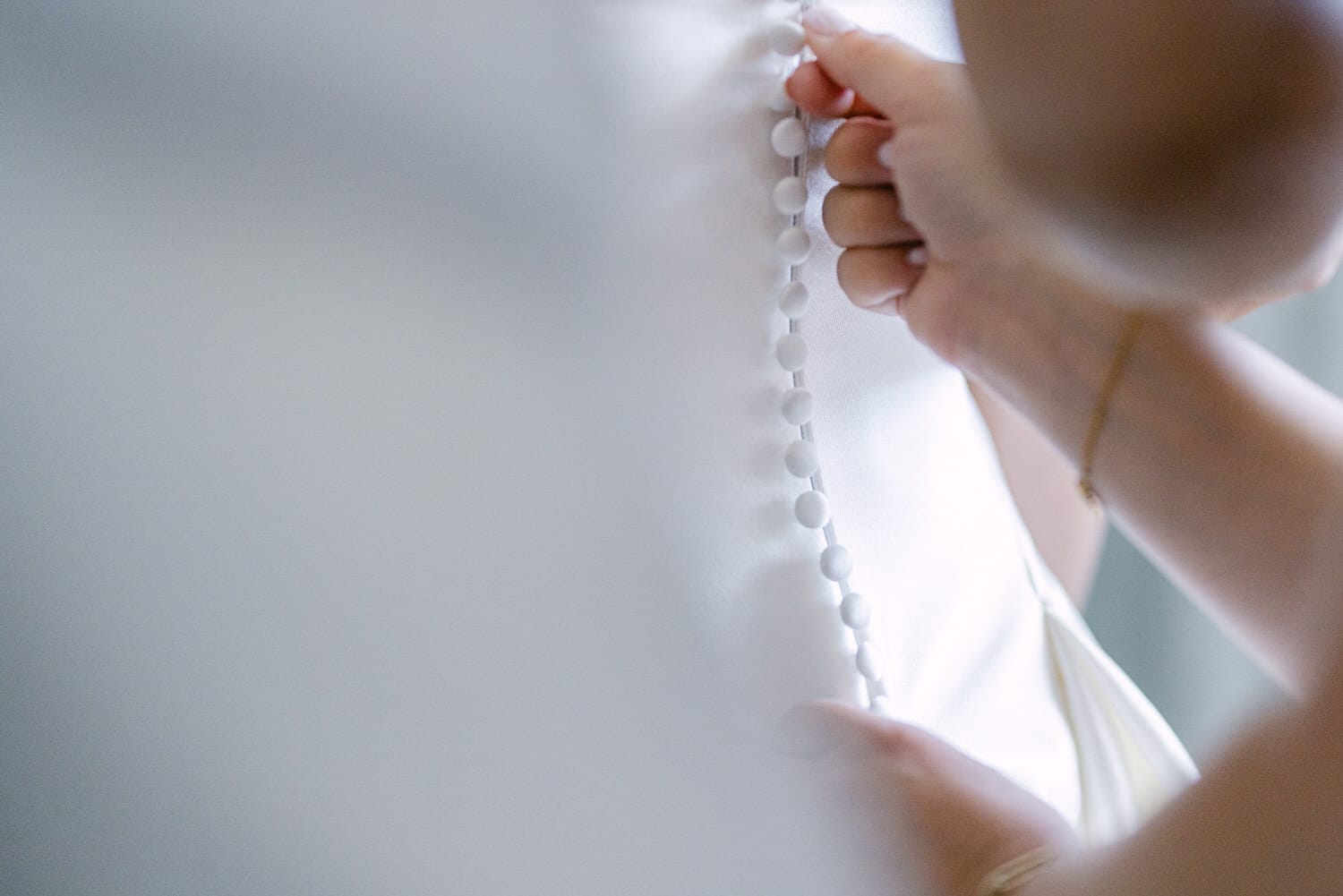 A close-up of hands buttoning a bridal gown, emphasizing the intricate details of the buttons against a soft, blurred background.