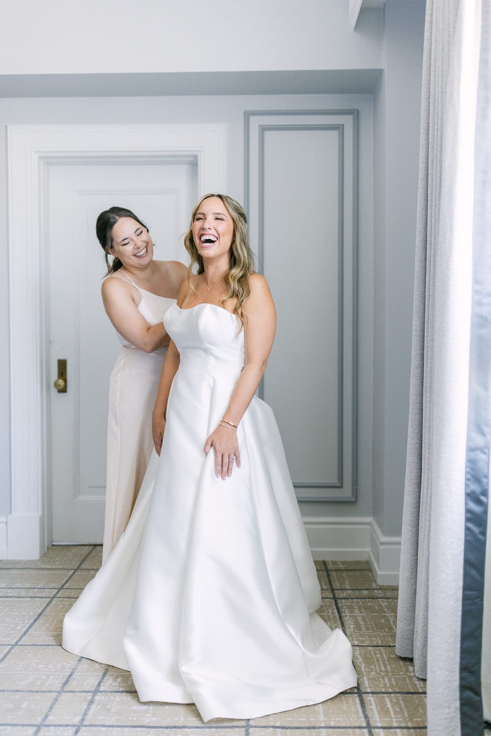 A bride laughing while her friend helps with her dress in a bright, elegant setting.