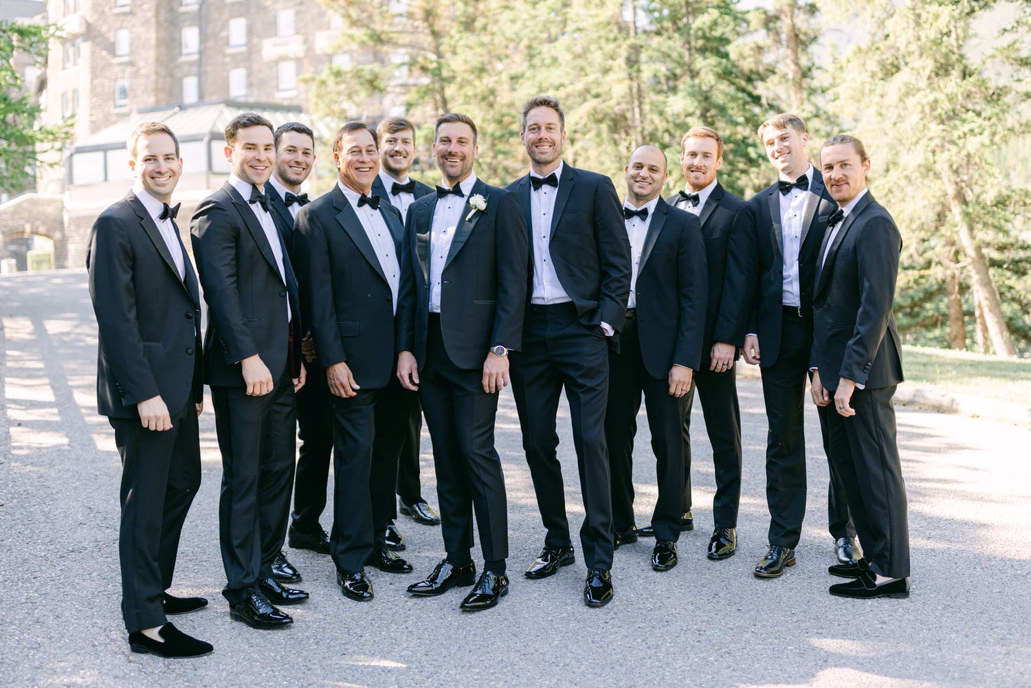 A cheerful group of groomsmen wearing black tuxedos and bow ties, posing together outdoors with a backdrop of trees and a stone building.