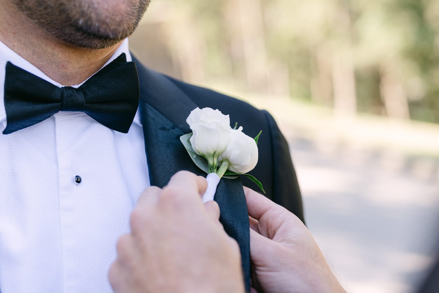 A close-up of a groom's tuxedo with a white boutonnière being pinned on his lapel.