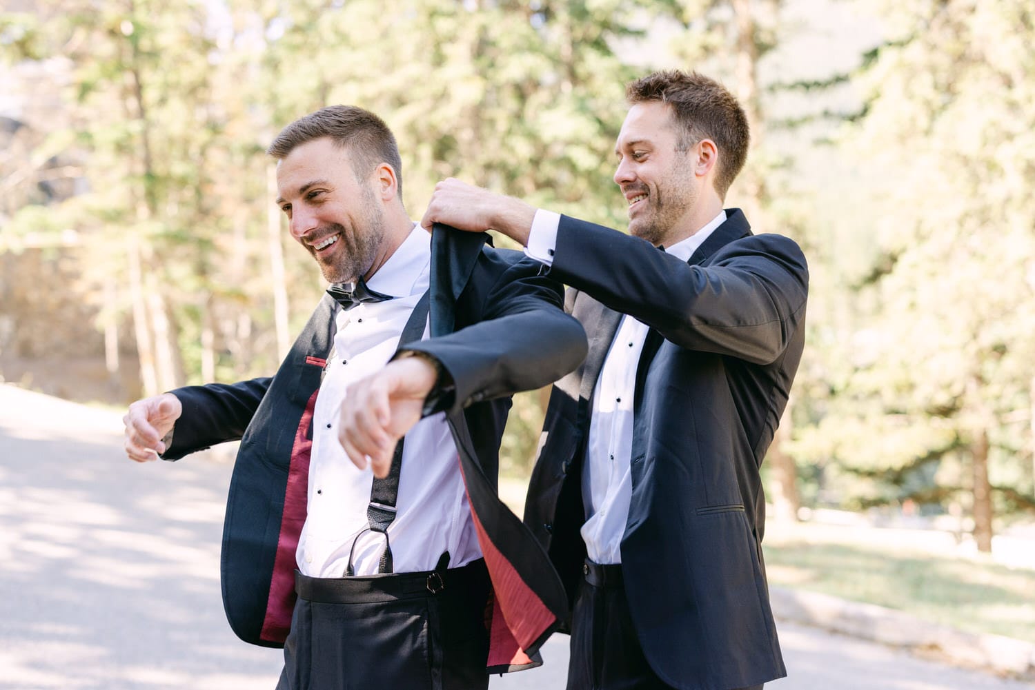 Two men in tuxedos joyfully assist each other in getting ready for a formal event outdoors surrounded by trees.