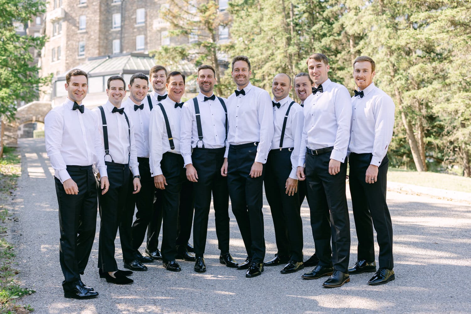 A group of ten men dressed in formal white shirts with bow ties and black pants, standing together on a path lined with greenery.