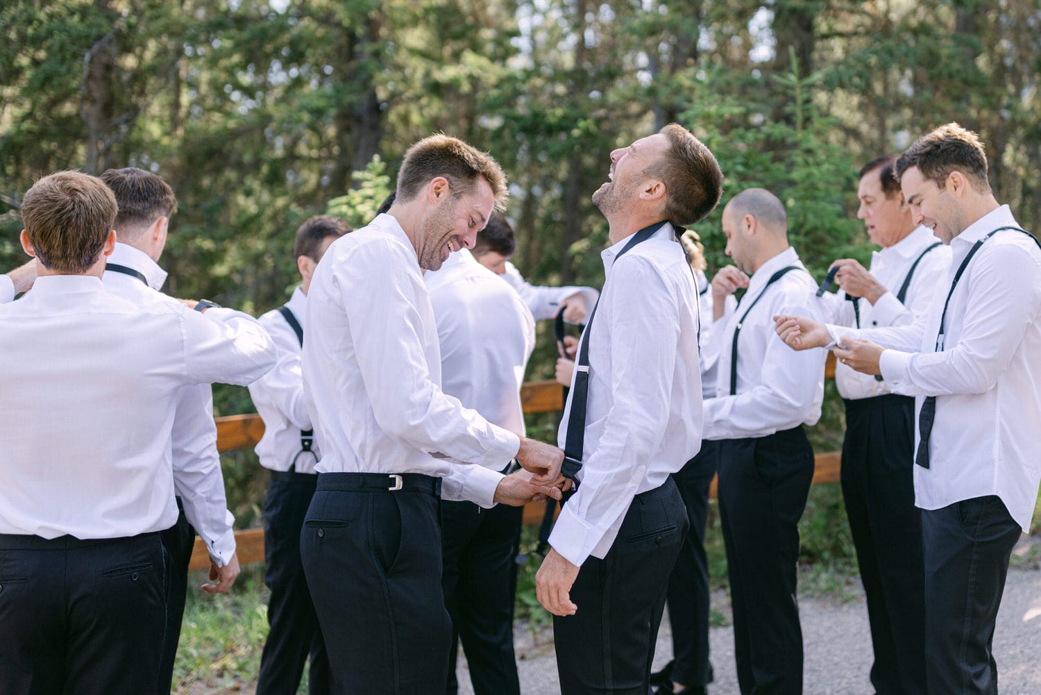 A group of groomsmen in white shirts and black pants share laughs and adjust their attire in a scenic outdoor setting.