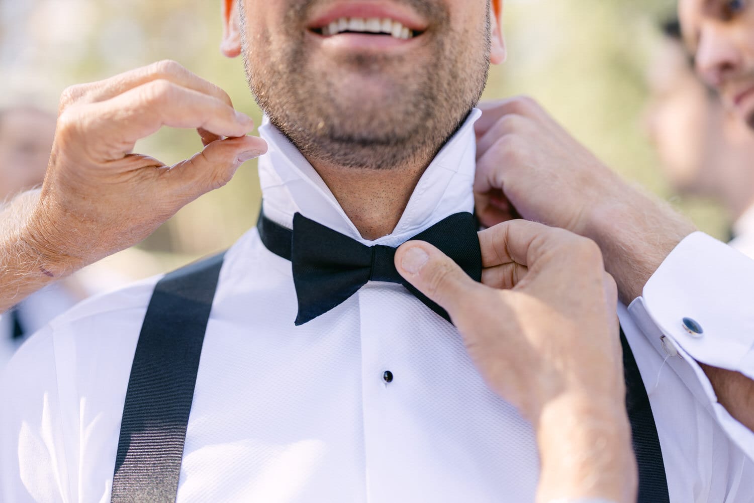 A groom adjusts his bow tie with help from friends, showcasing the details of his formal attire and the camaraderie of his wedding preparations.