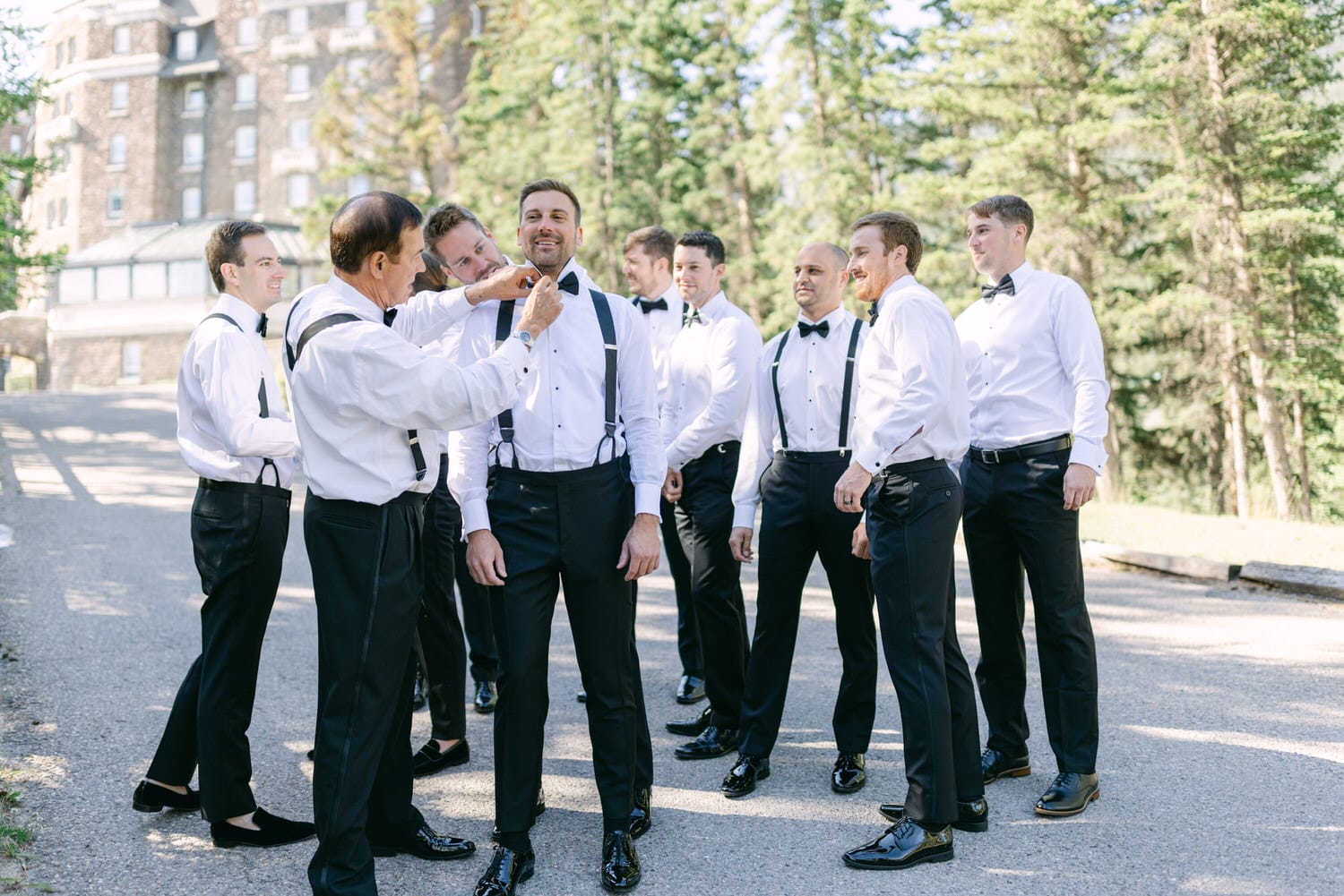 A group of groomsmen in formal attire, adjusting clothing and bowties outdoors before a wedding, surrounded by lush greenery and a charming venue in the background.