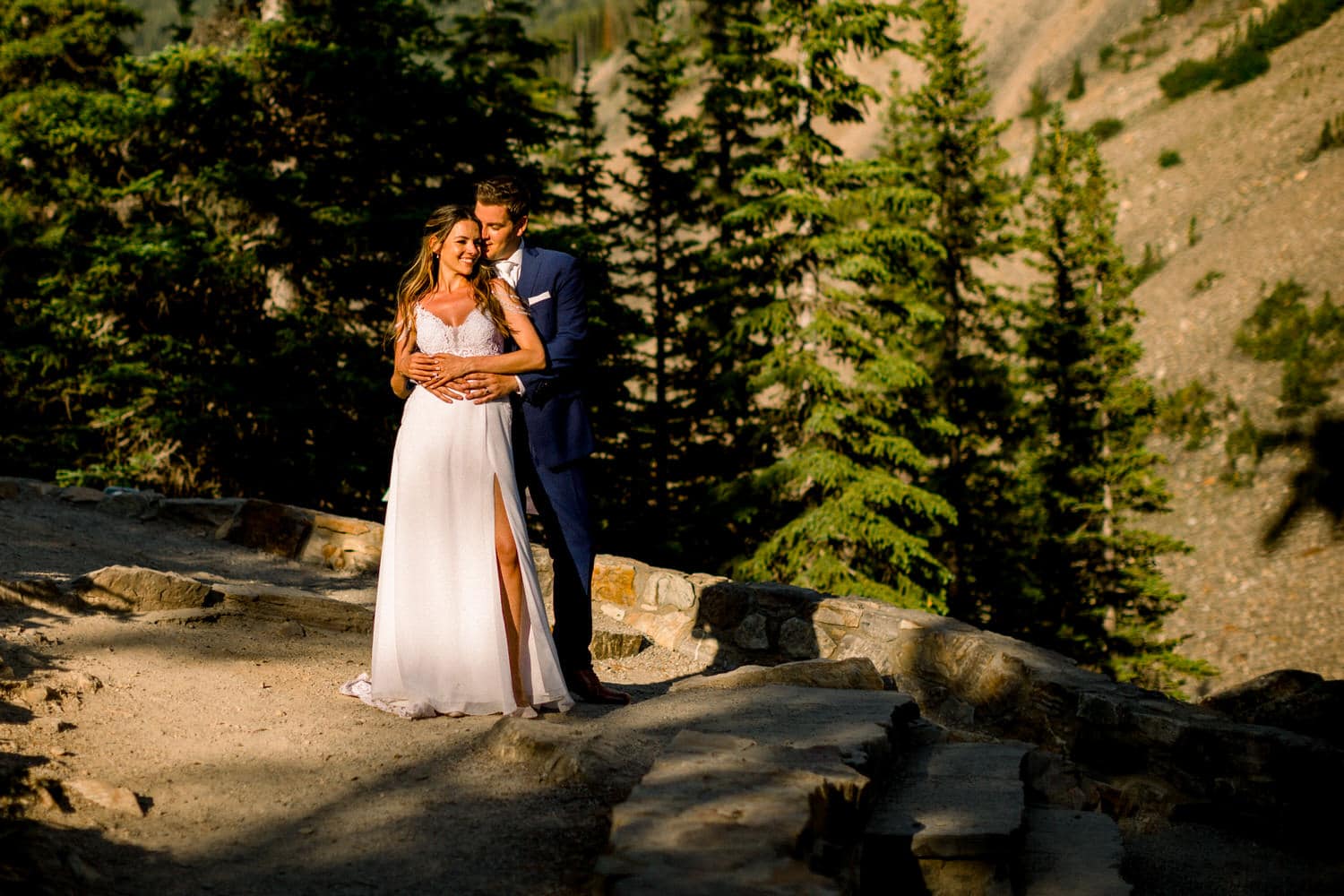 A couple in formal attire stands together on a scenic path, surrounded by tall pine trees, capturing a moment of love and intimacy.