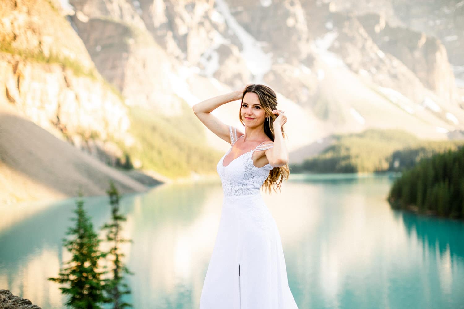 A woman in a white wedding dress poses gracefully by a serene turquoise lake surrounded by mountains and greenery.