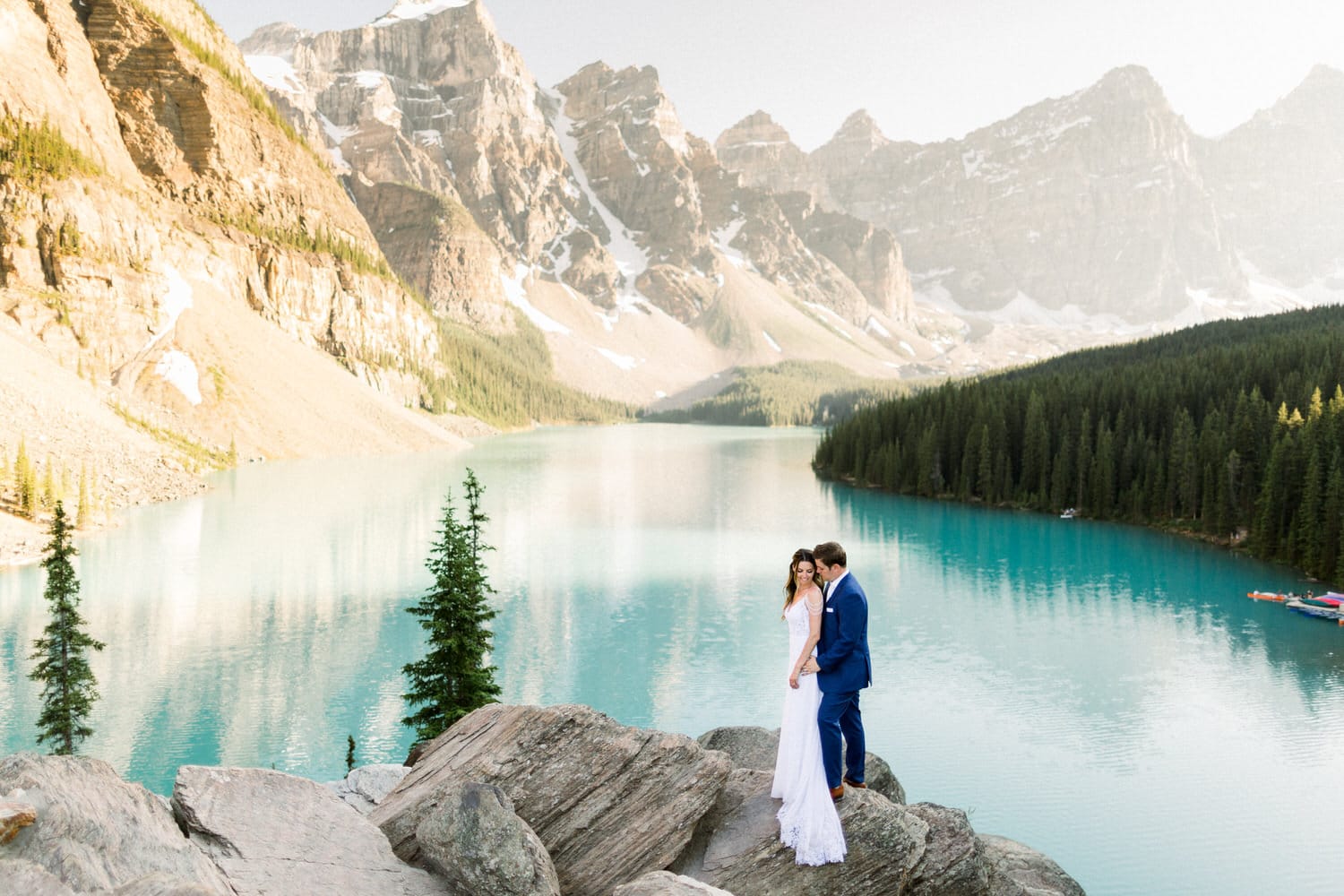 A couple in elegant wedding attire stands on rocky terrain by a serene turquoise lake, surrounded by majestic mountains and lush forests, capturing a moment of intimacy amidst stunning natural scenery.