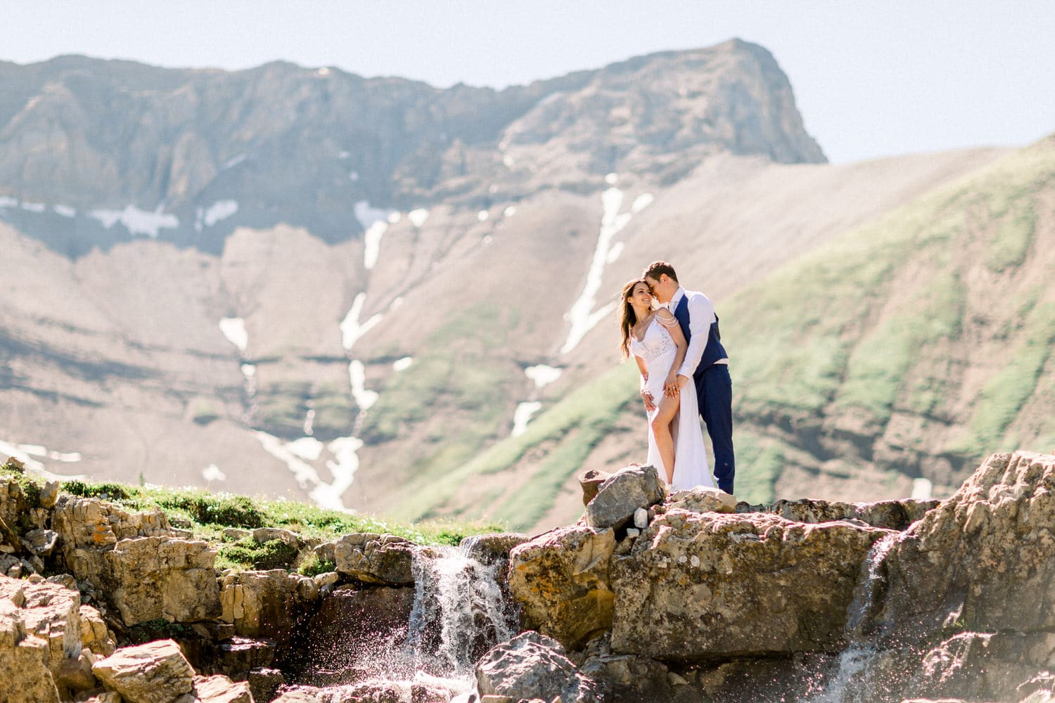 A couple in wedding attire stands on a rocky ledge near a small waterfall, surrounded by lush green mountains and clear blue sky, sharing a tender moment.